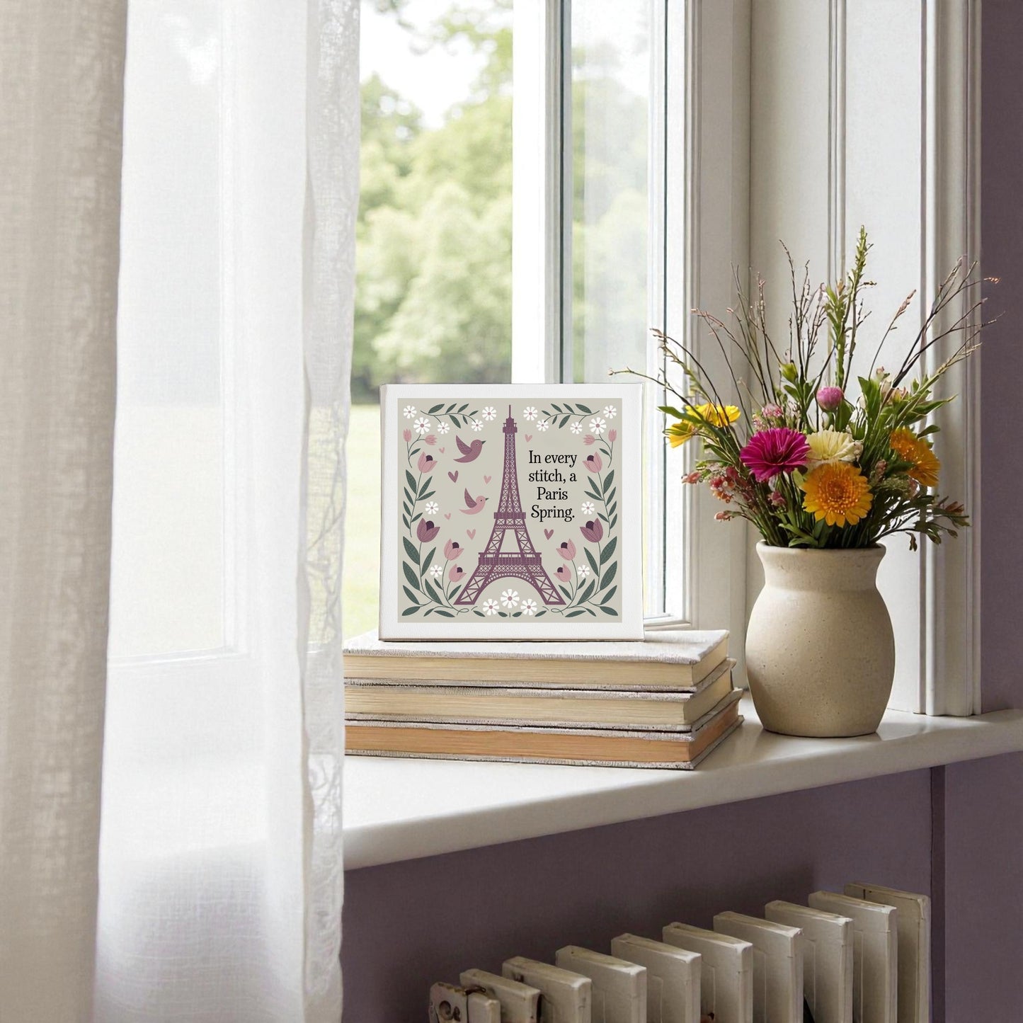 A vase of colorful flowers rests on books by a window, beside Mirabilia Boutique's Paris Spring Stitch Canvas—embroidered-style floral art with birds and blooms—evoking cottagecore Paris vibes as sunlight filters through sheer curtains.