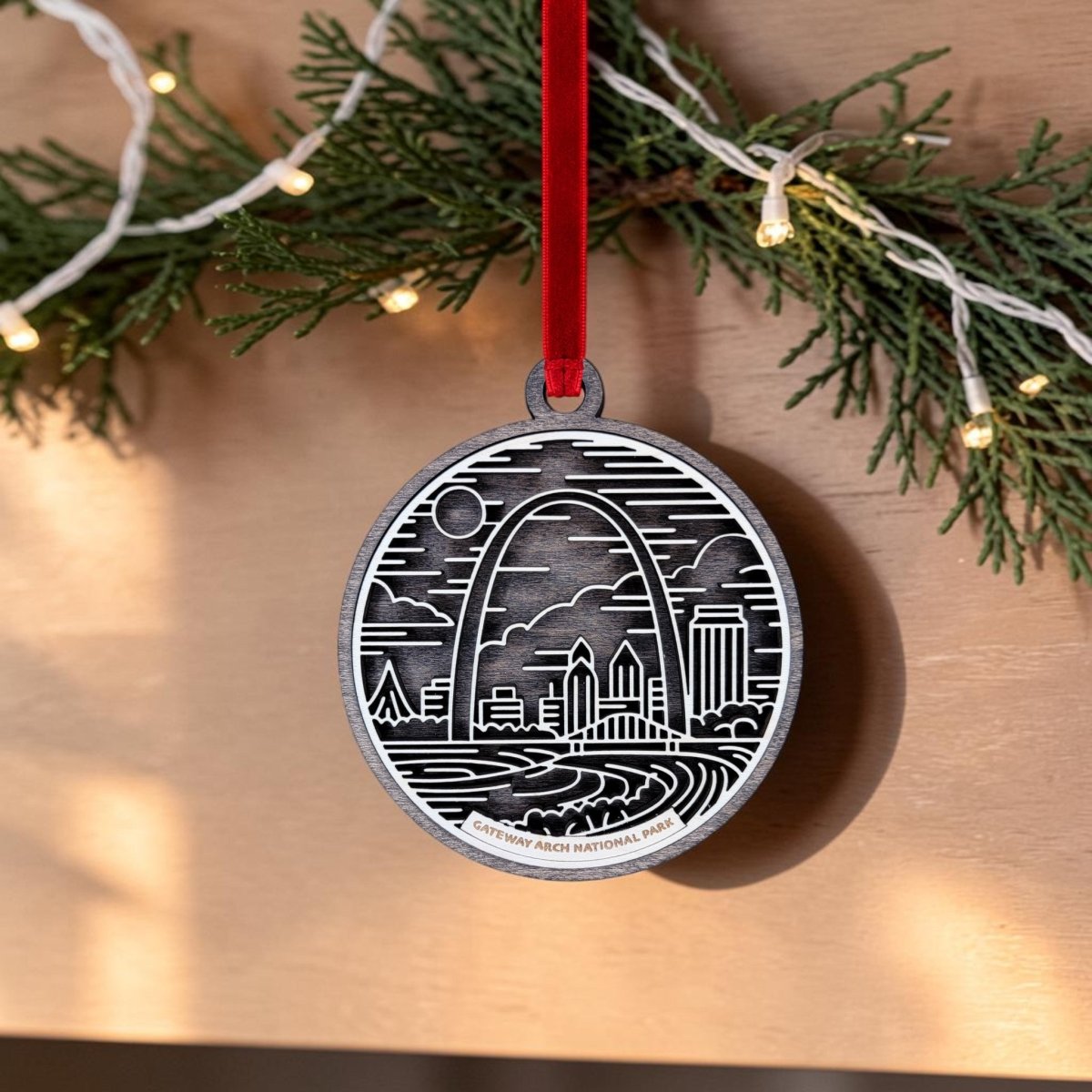 The Gateway Arch National Park Line Art Ornament by Mirabilia Boutique features an engraved line art design of the Gateway Arch and cityscape, hanging from a red ribbon amidst pine garland and string lights—making it the perfect St. Louis souvenir for any holiday celebration.