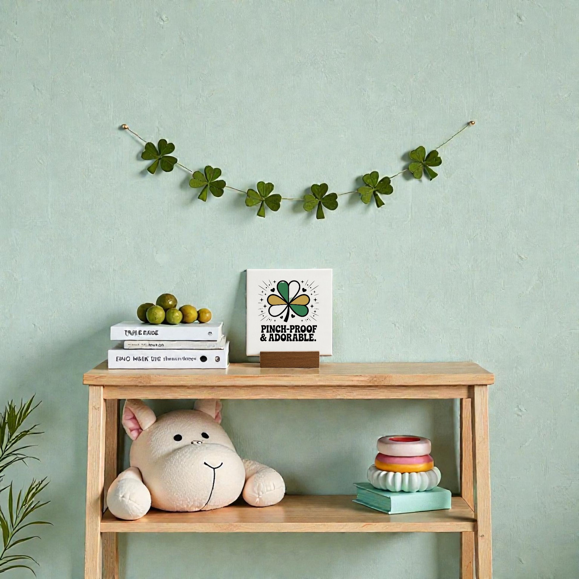 A wooden shelf displays Mirabilia Boutique’s Pinch-Proof & Adorable Canvas Sign (4x4), plus a plush hippo, stacked rings, and books. Overhead, a green four-leaf clover garland adds festive St. Patrick’s Day flair to the pale green kids room.