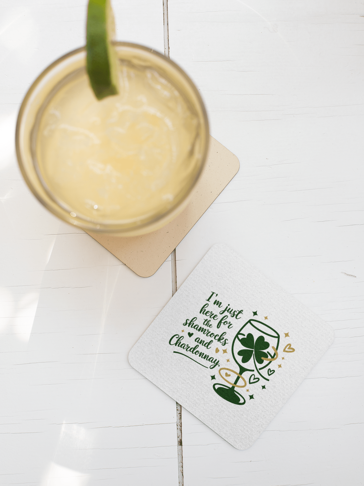 A glass of light yellow drink with ice and lime rests beside a Mirabilia Boutique Shamrocks and Chardonnay Coaster from their set—a festive piece adorned with shamrocks, wine glasses, and the saying "I'm just here for the shenanigans and chardonnay.