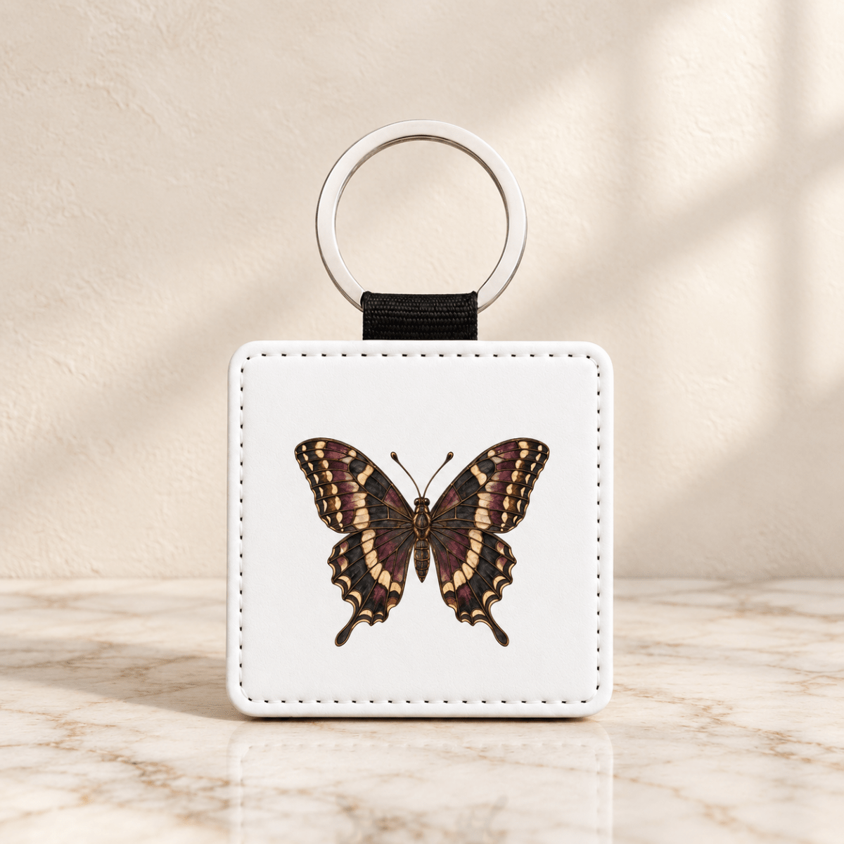 The Stained Glass Swallowtail Keychain from Mirabilia Boutique’s Velvet Ruins collection features a detailed brown and yellow butterfly illustration on a square white base with a metal ring. This elegant butterfly gift is displayed atop a marble surface, bathed in soft sunlight.