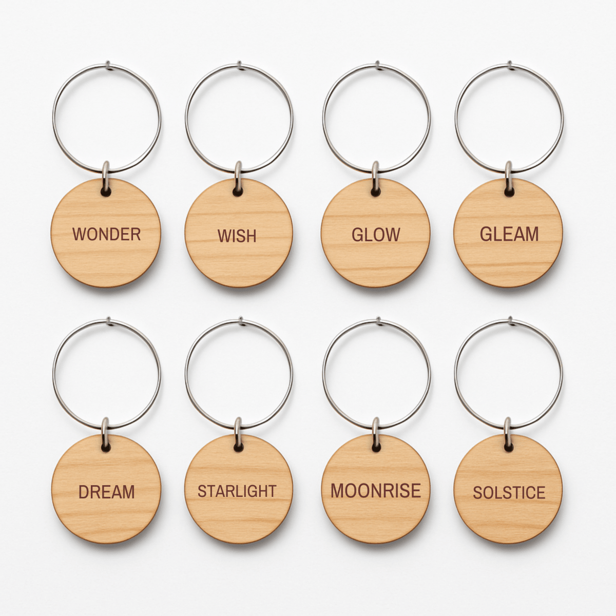 Eight round wooden wine charms by Mirabilia Boutique, from the Star & Solstice Engraved Wood Wine Charms Set of 8, are displayed in two rows with metal rings. Each charm features a different engraved word: Wonder, Wish, Glow, Gleam, Dream, Starlight, Moonrise, and Solstice. These unique pieces can be used as celestial or personalized laser-engraved wine charms to add a magical touch to any gathering.