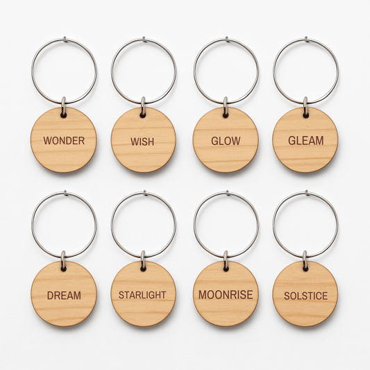 Eight round wooden wine charms by Mirabilia Boutique, from the Star & Solstice Engraved Wood Wine Charms Set of 8, are displayed in two rows with metal rings. Each charm features a different engraved word: Wonder, Wish, Glow, Gleam, Dream, Starlight, Moonrise, and Solstice. These unique pieces can be used as celestial or personalized laser-engraved wine charms to add a magical touch to any gathering.