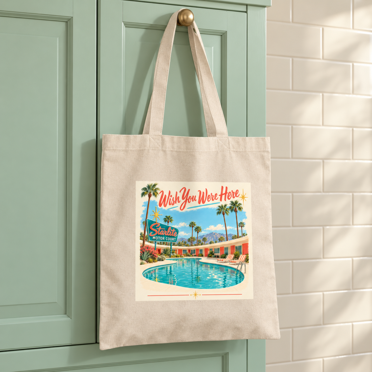Mirabilia Boutique Wish You Were Here Tote hanging from a cabinet knob, featuring a mid-century postcard graphic of a motel swimming pool under palm trees with the text “Wish You Were Here” and “Starlite Motor Court.”.