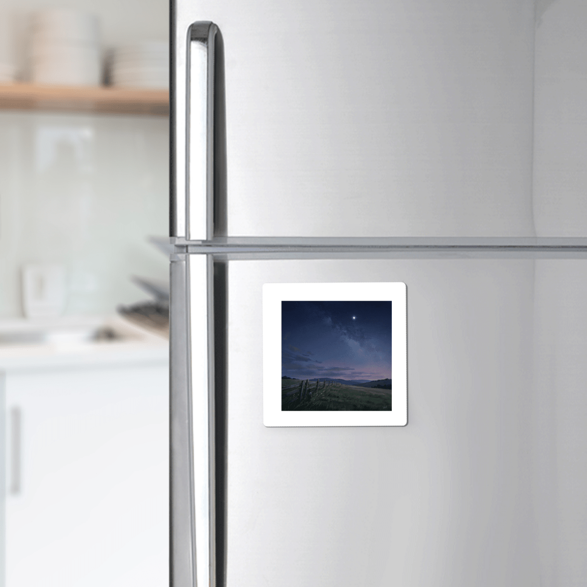 The Stay For The Stars Magnet by Mirabilia Boutique, featuring a twilight countryside evening landscape, is displayed on a stainless steel refrigerator door in a modern kitchen.