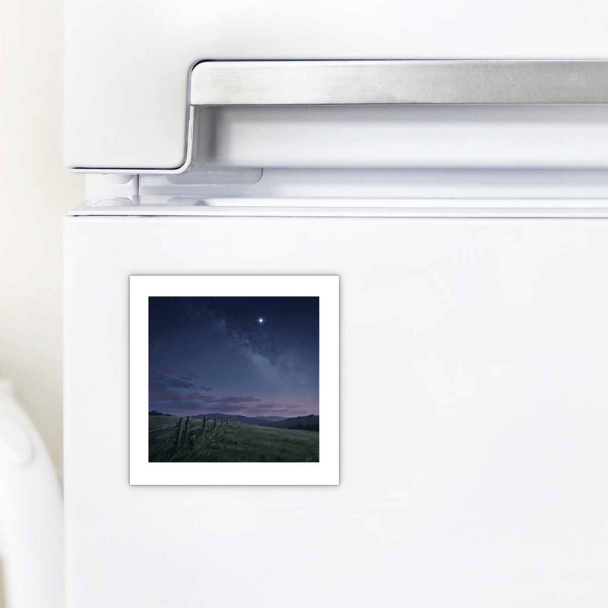 The Stay For The Stars Magnet by Mirabilia Boutique, showcasing a twilight landscape with a starry night sky and grassy field, is displayed on a white refrigerator door beside the silver handle.