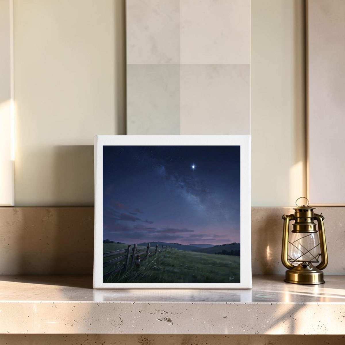 An 8x8 Stay For The Stars Canvas from Mirabilia Boutique rests on a light-colored counter, showcasing a tranquil night landscape with a bright star shining above gentle fields and a wooden fence—perfect for country meadow décor. To the right of the canvas, an antique-style brass lantern adds a charming, vintage touch.