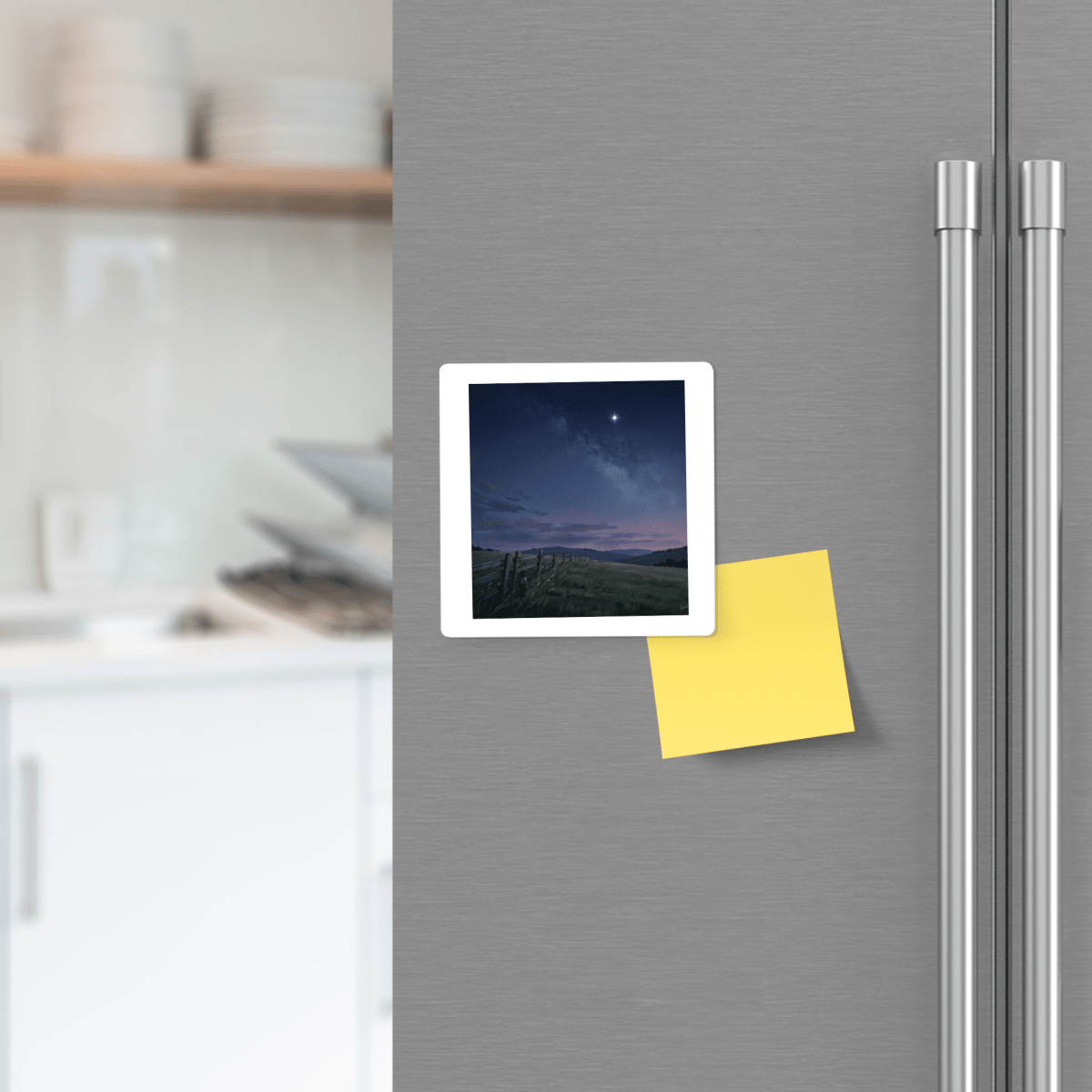 A “Stay For The Stars Magnet” by Mirabilia Boutique, featuring a photo of a night sky and a yellow sticky note, is attached to a stainless steel refrigerator door in a modern kitchen.
