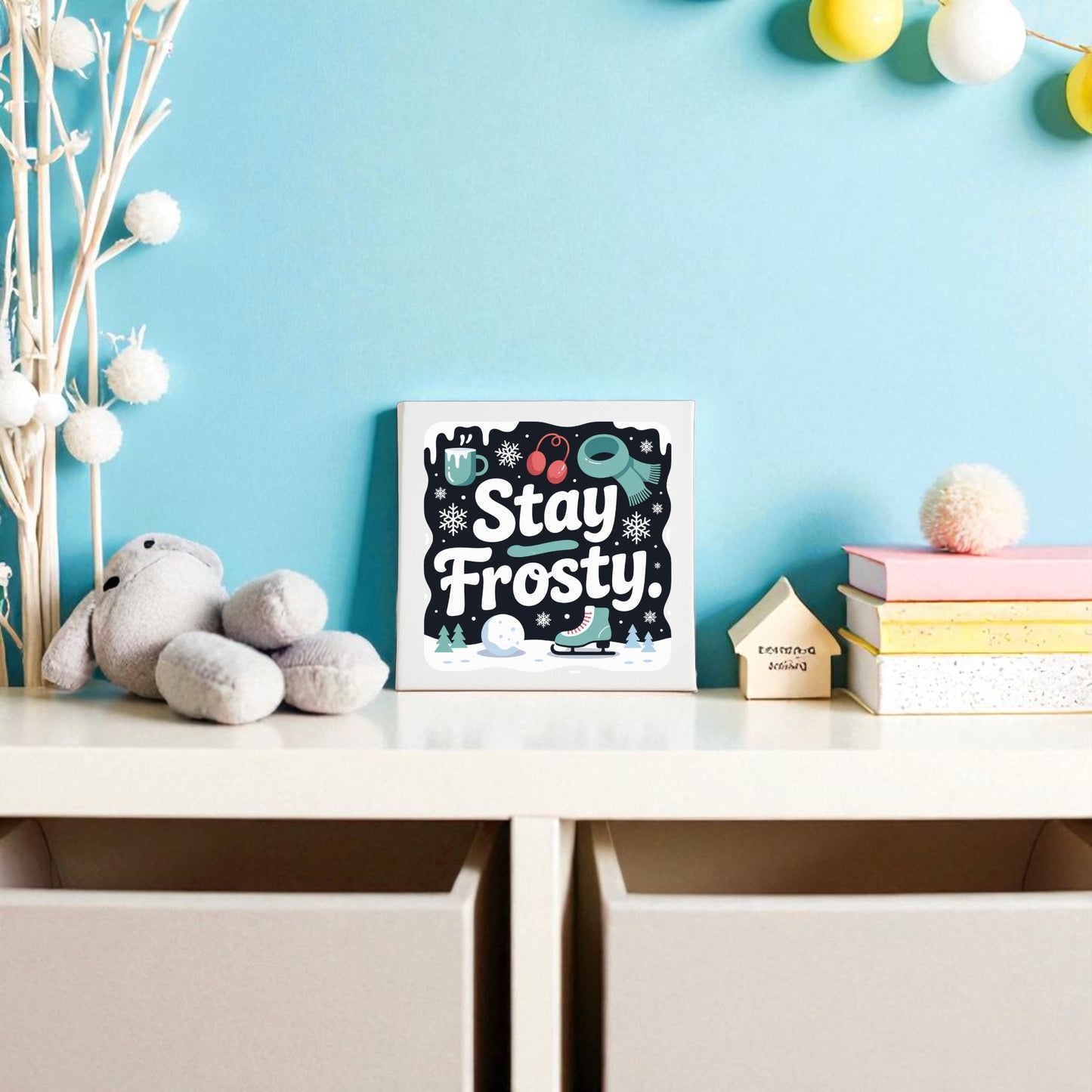 A nursery shelf displays Mirabilia Boutique's “Stay Frosty” canvas sign (8x8 winter wall art), a stuffed rabbit, books, and wooden house decor against a light blue wall with string lights and pom-pom winter decorations.