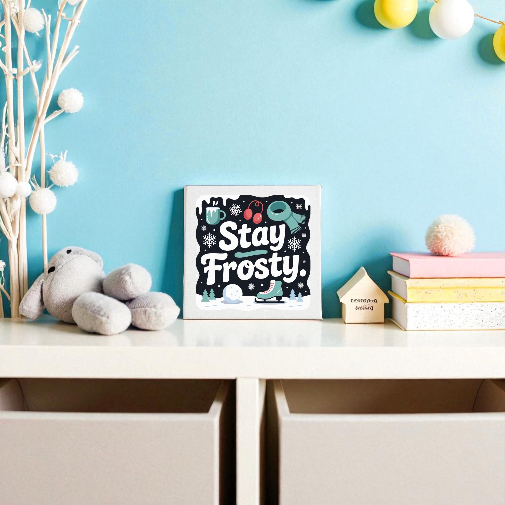 A nursery shelf displays Mirabilia Boutique's “Stay Frosty” canvas sign (8x8 winter wall art), a stuffed rabbit, books, and wooden house decor against a light blue wall with string lights and pom-pom winter decorations.