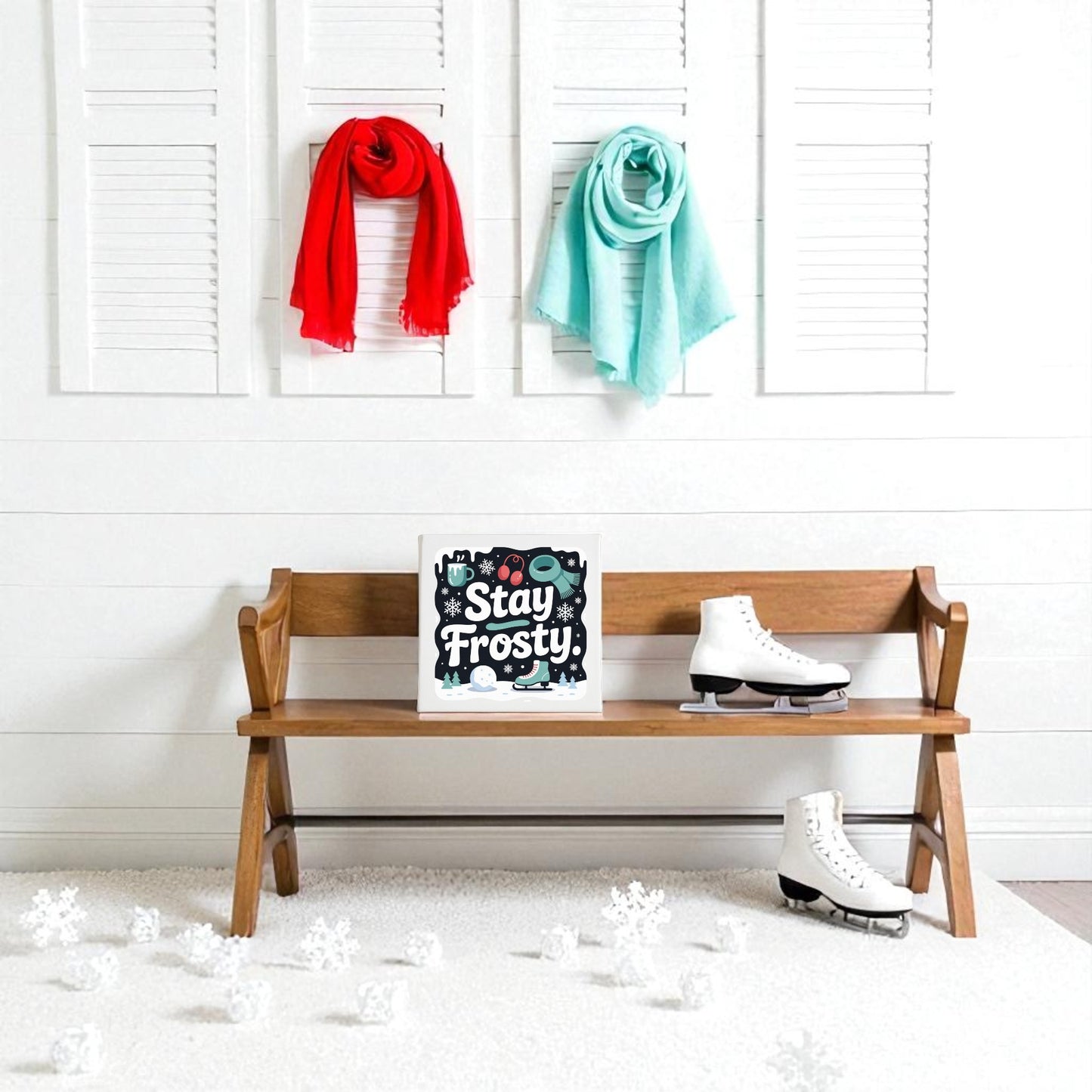 A wooden bench displays Mirabilia Boutique's "Stay Frosty" Canvas Sign (8x8 Winter Wall Art) and an ice skate, with another skate on the white carpet. Red and teal scarves hang above, and snowflake winter decor is scattered on the floor.