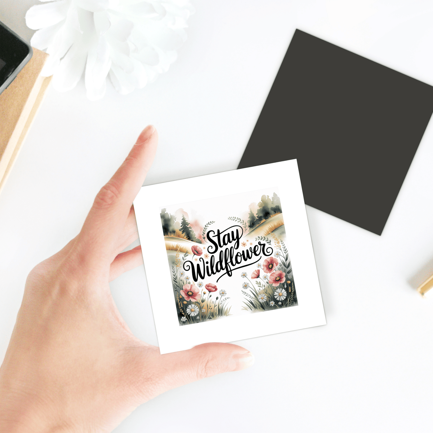 A hand holds the "Stay Wildflower Magnet" by Mirabilia Boutique, featuring a floral quote design—ideal for decorating your fridge or locker. A black card, white flower, and part of a notebook rest nearby on a white surface.