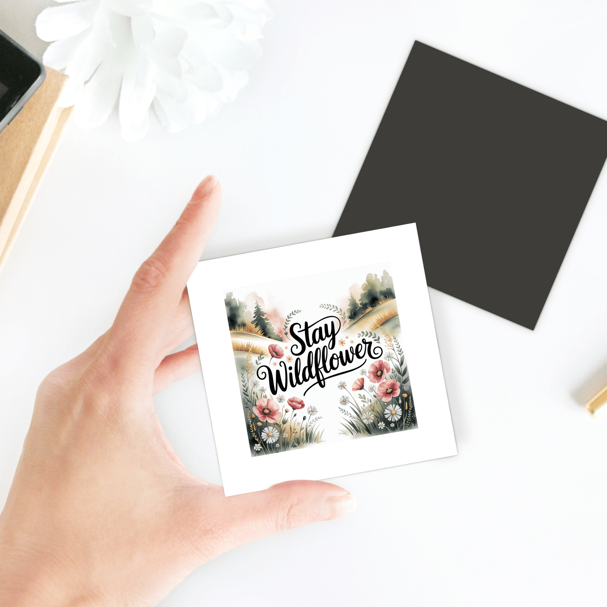 A hand holds the "Stay Wildflower Magnet" by Mirabilia Boutique, featuring a floral quote design—ideal for decorating your fridge or locker. A black card, white flower, and part of a notebook rest nearby on a white surface.