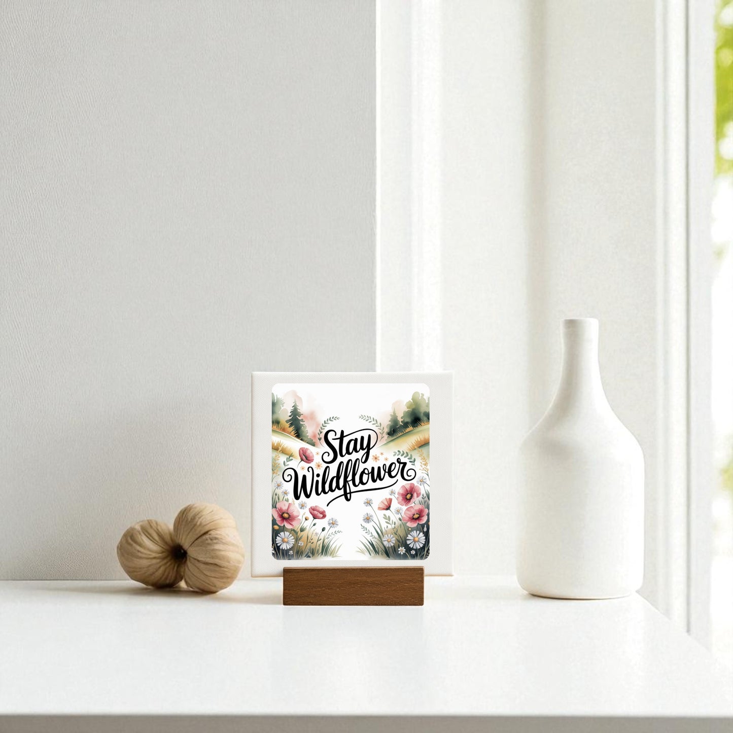 A minimal white table holds a Mirabilia Boutique Stay Wildflower Mini Sign with floral decor, a decorative white vase, and a tan accent piece, all arranged near a sunlit window.