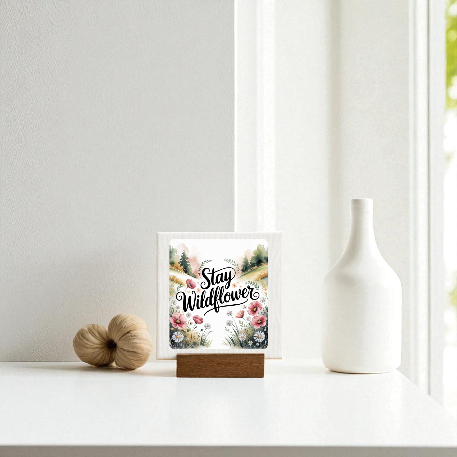 A minimal white table holds a Mirabilia Boutique Stay Wildflower Mini Sign with floral decor, a decorative white vase, and a tan accent piece, all arranged near a sunlit window.