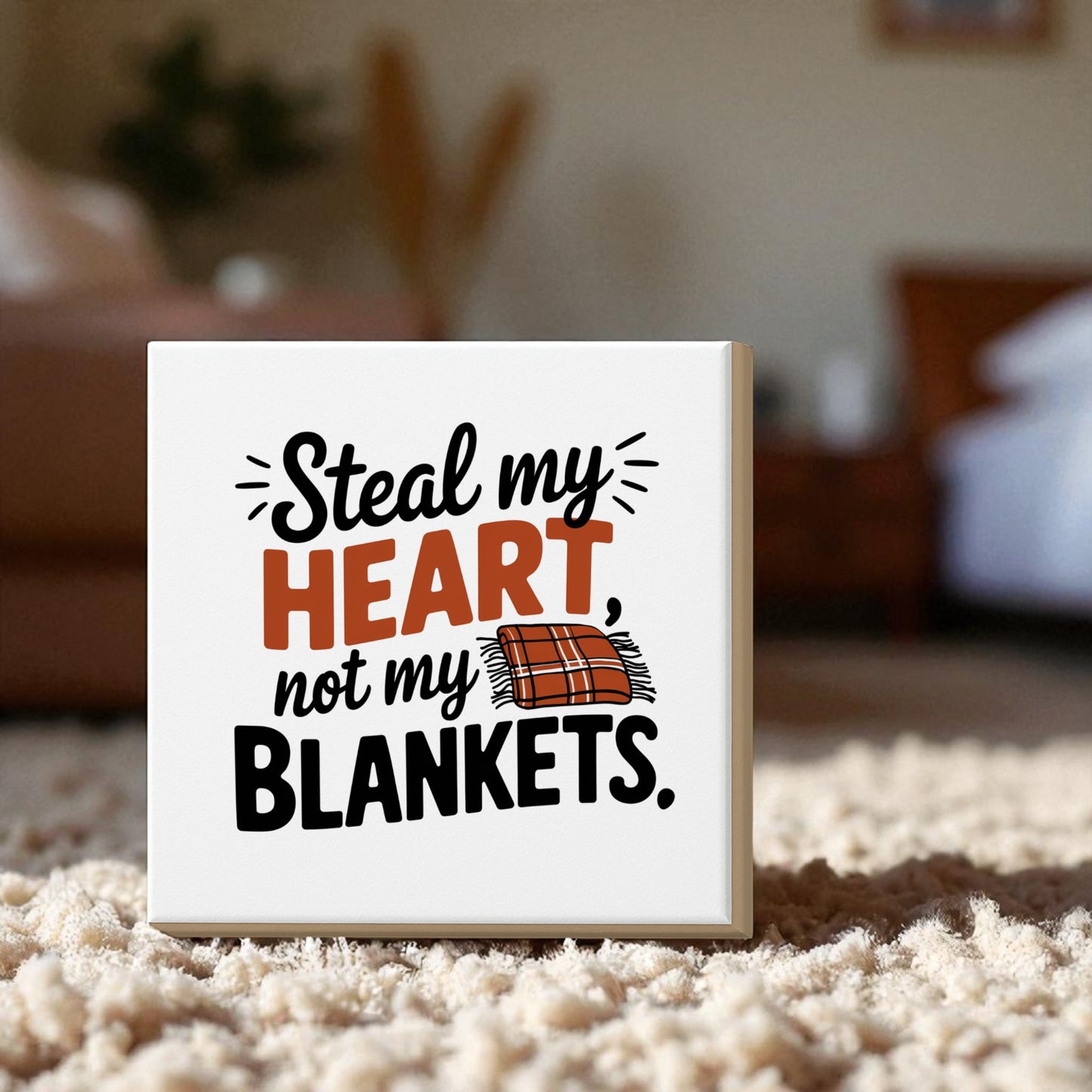 The Mirabilia Boutique "Steal My Heart Not My Blankets" 8x8 canvas sign features “blankets” with a plaid blanket graphic—funny bedroom décor and a cute couple’s gift, displayed on a carpeted floor with a blurred bedroom backdrop.