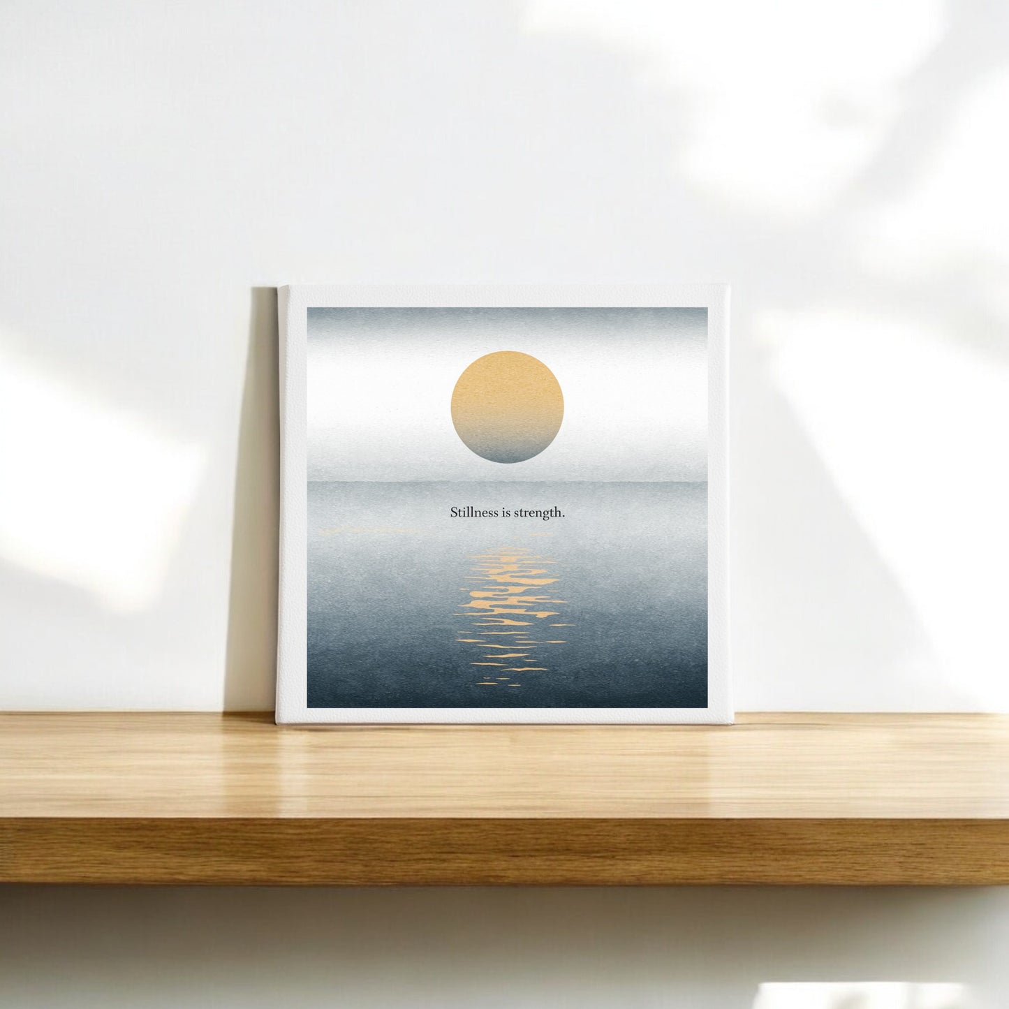 The Stillness Is Strength Canvas by Mirabilia Boutique is a minimalist square wall art featuring a sun over calm water with its reflection and the quote “Stillness is strength.”—a serene choice for meditation decor.