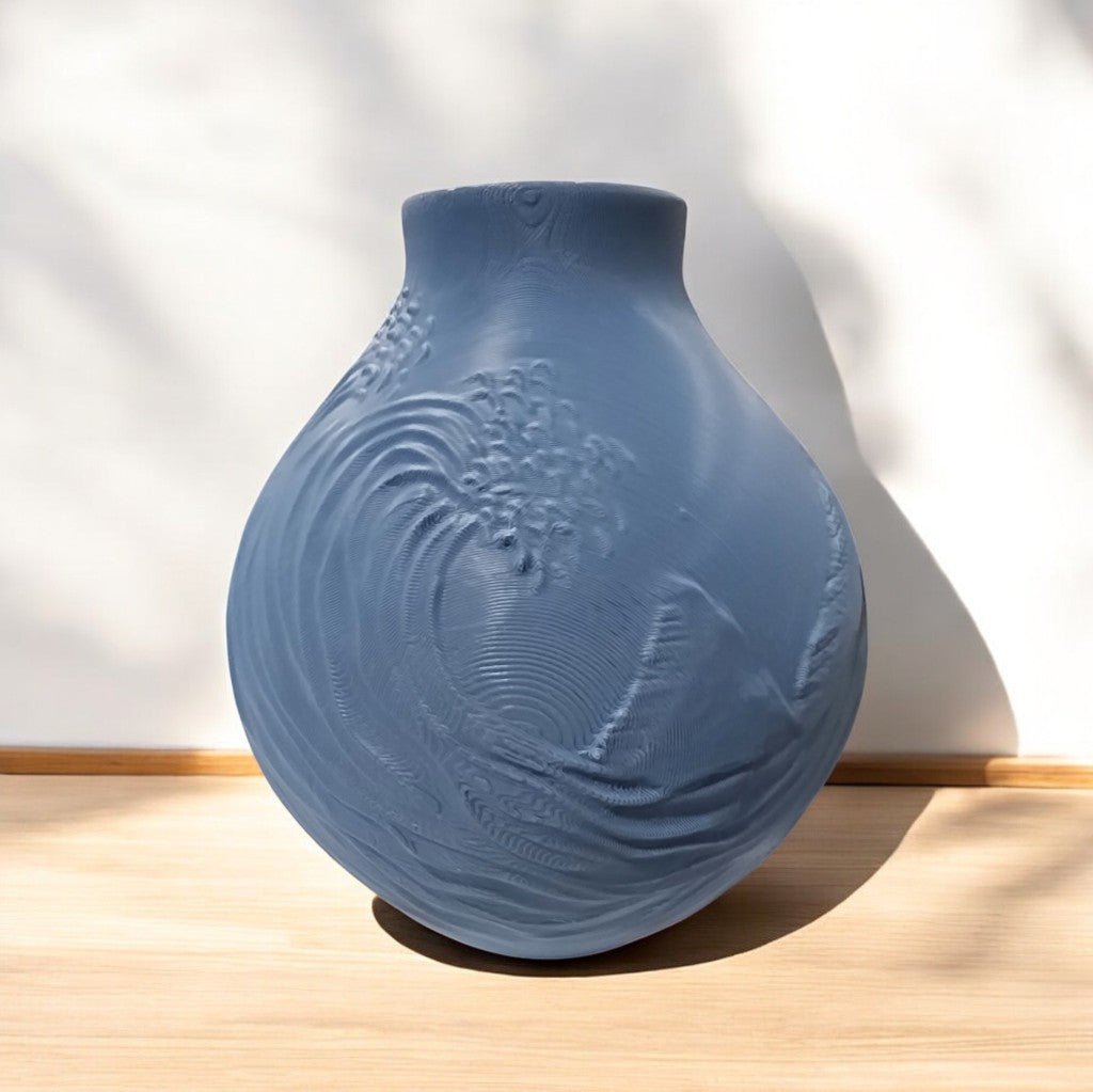 The Stone Blue Coastal Wave Vase by Mirabilia Boutique features a round, matte finish and wave-like embossing. It sits on a light wooden surface against a white background with soft shadows.