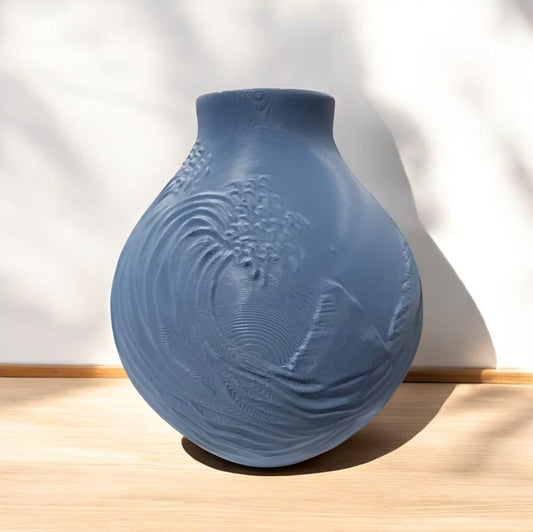 The Stone Blue Coastal Wave Vase by Mirabilia Boutique features a round, matte finish and wave-like embossing. It sits on a light wooden surface against a white background with soft shadows.