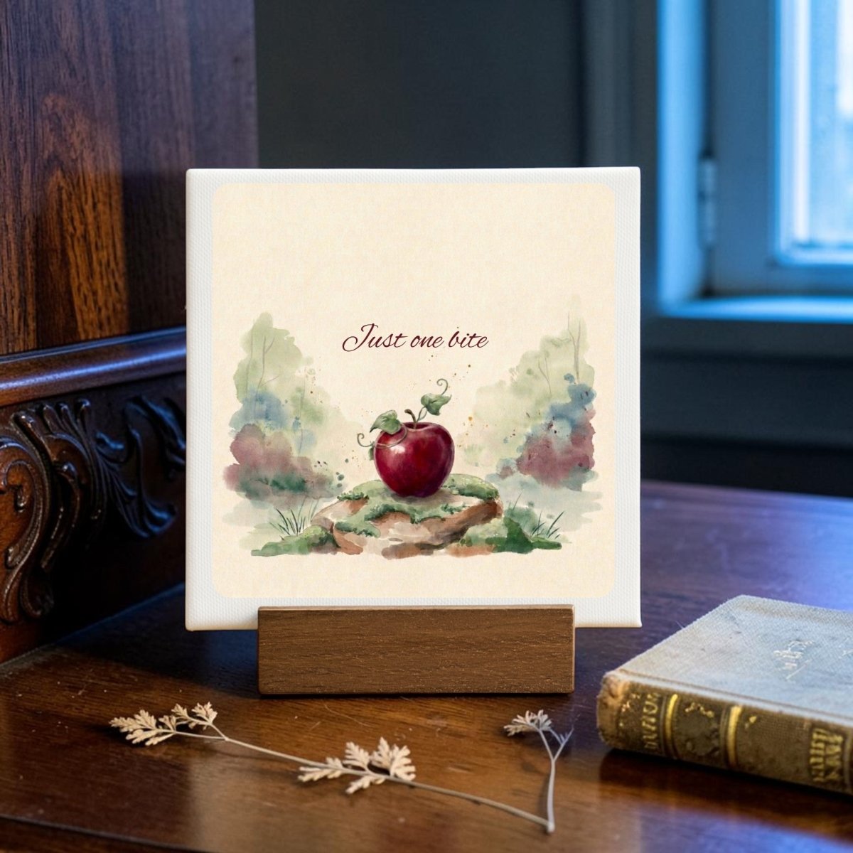 The "Just One Bite Mini Canvas Sign" by Mirabilia Boutique features a painting of a red apple and the words "Just one bite." Displayed on a wooden stand atop a desk near a closed book and dried flowers with a window in the background, this charming accent brings fairy tale decor to any space.