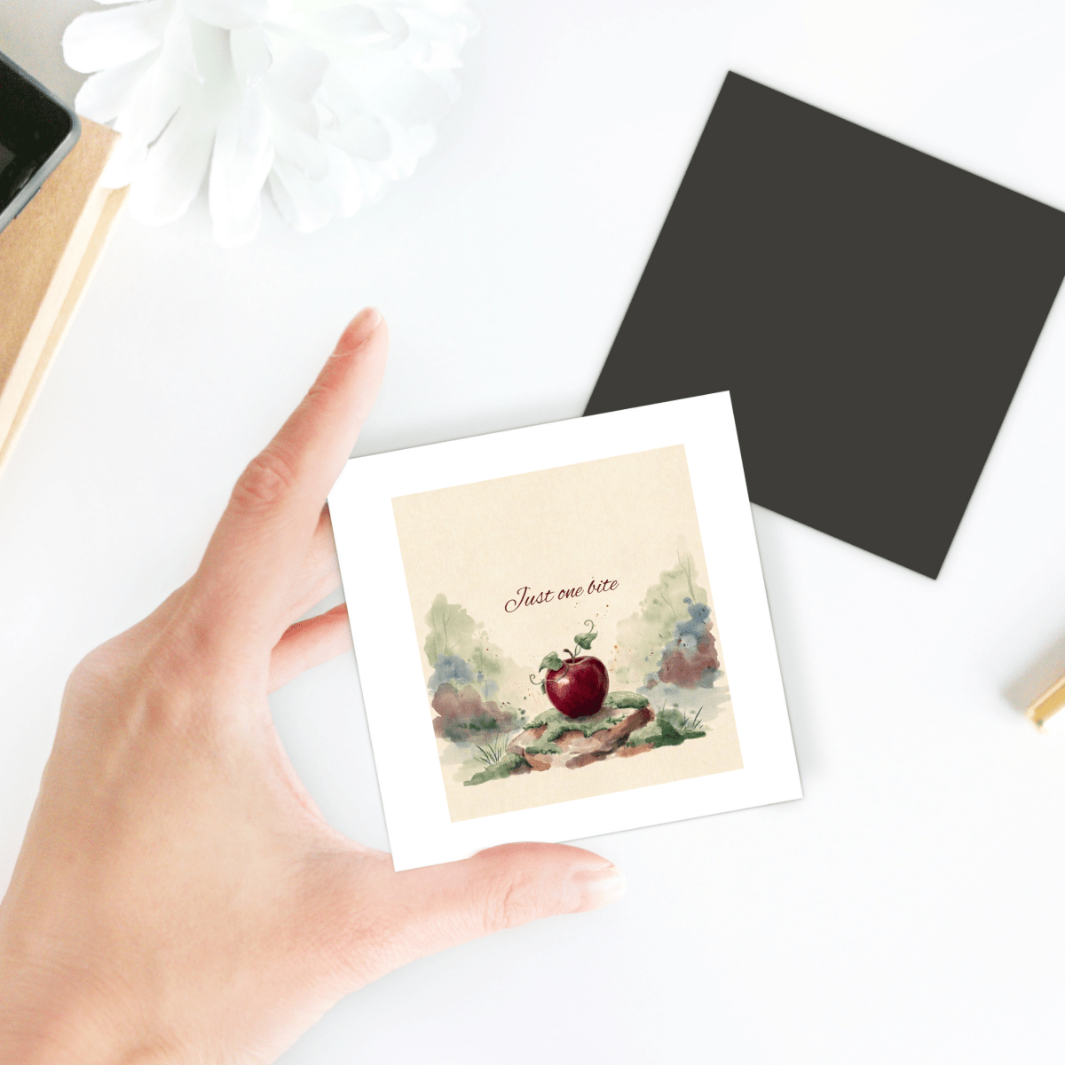 A hand holds the Just One Bite Fairy Tale Apple Magnet by Mirabilia Boutique, featuring a watercolor apple and the phrase “Just one bite.” Nearby, a black square card, a white flower, and various desk items rest on a white surface, enhancing the charming cottagecore décor.