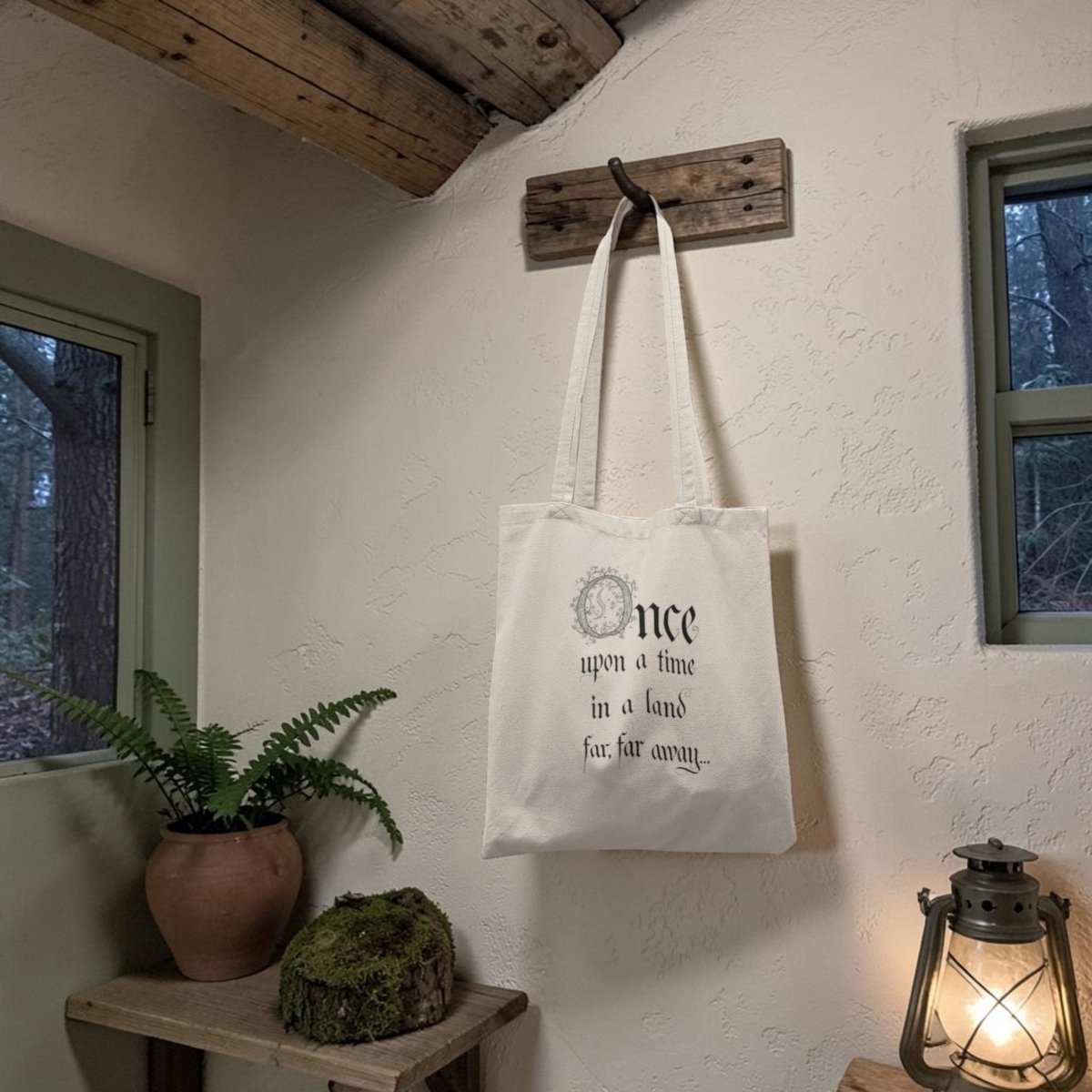 The Once Upon a Time Fairytale Tote Bag by Mirabilia Boutique, featuring the phrase “Once upon a time in a land far, far away…”, hangs from a wooden hook in a rustic room beside a potted fern, lush moss, an antique lantern, and a window overlooking the trees.