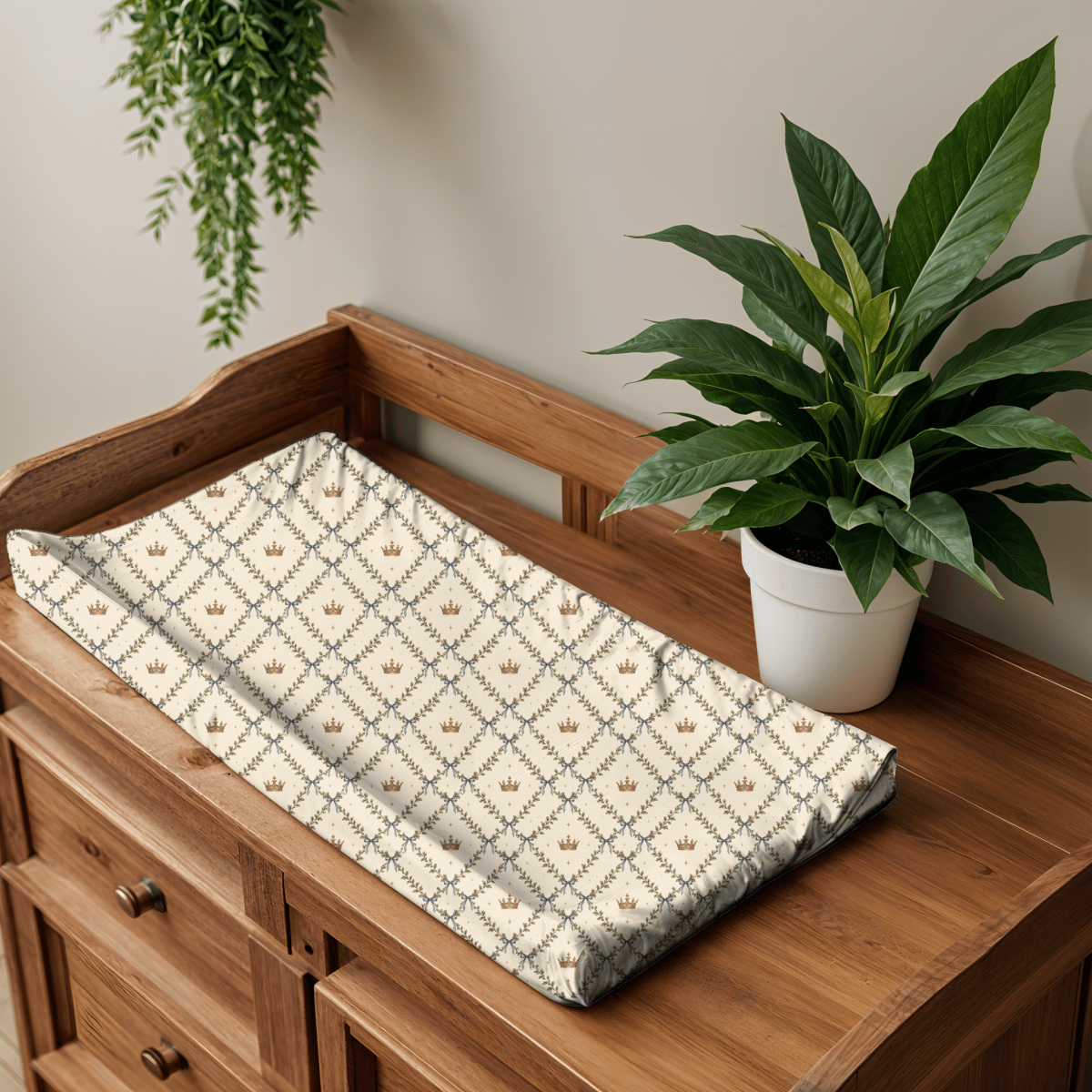 The Storybook Trellis Changing Pad Cover from Mirabilia Boutique’s Moon & Moss Nursery collection sits atop a wooden dresser next to a potted green plant. Its cream-colored, jersey knit polyester fabric showcases a charming repeating diamond motif with olive branches, blue ribbons, and petite gold crowns. Ideal for woodland nursery decor, this enchanting changing pad cover is complemented by the presence of a hanging plant in the upper left corner.