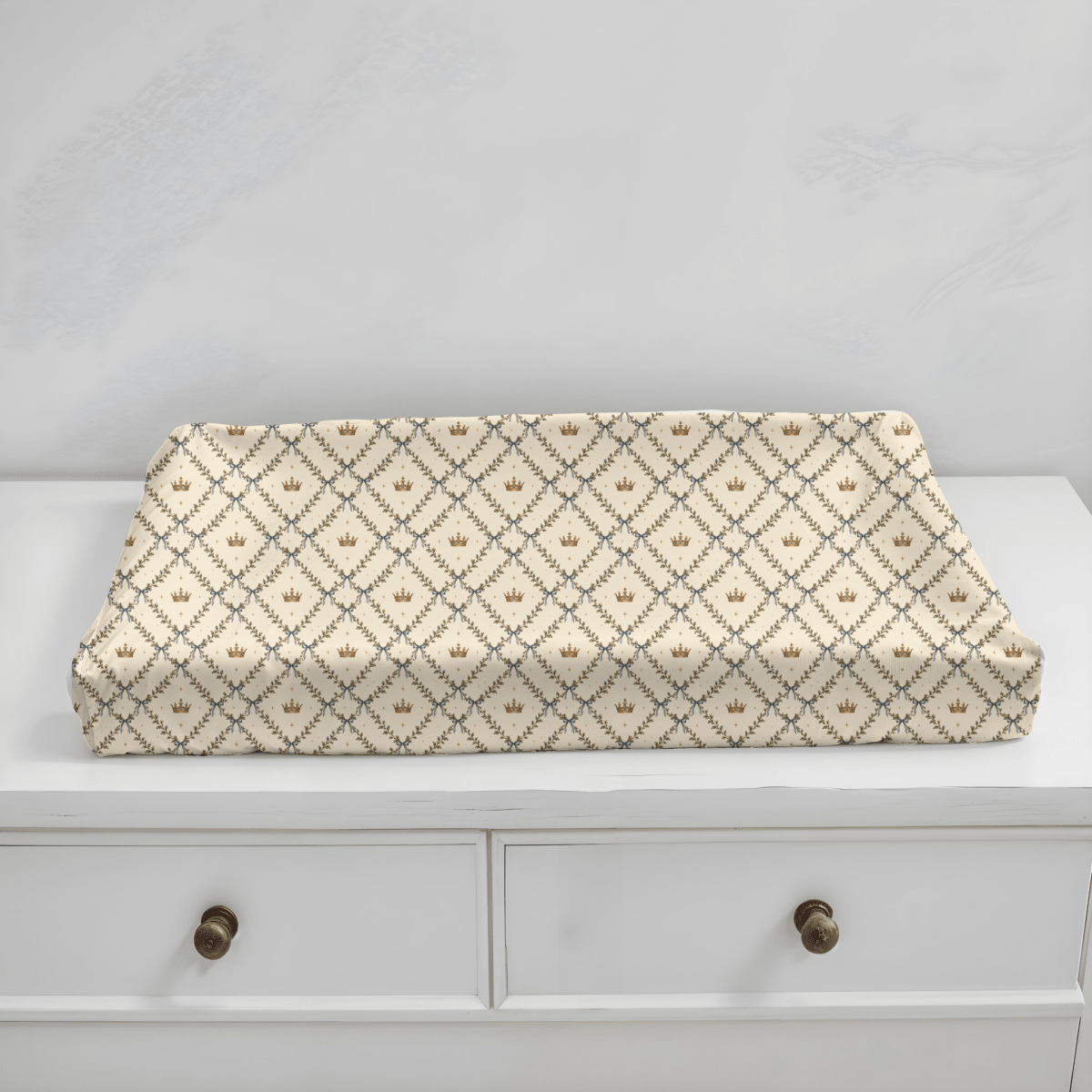 The Storybook Trellis Changing Pad Cover from Mirabilia Boutique’s Moon & Moss Nursery collection rests atop a white, two-drawer dresser with round wooden knobs, its cream-colored jersey knit surface adorned with a brown and gray geometric crown pattern for a cozy woodland nursery accent.