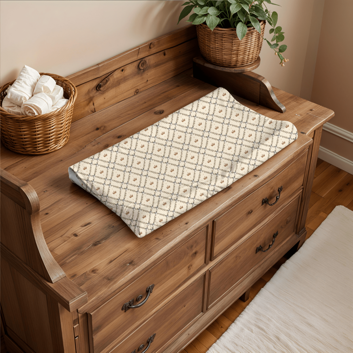 A wooden changing table with the Mirabilia Boutique Storybook Trellis Changing Pad Cover from the Moon & Moss Nursery collection, crafted from soft jersey knit polyester, paired with a basket of rolled towels, a potted plant, and a white rug on a hardwood floor creates the perfect woodland nursery decor.