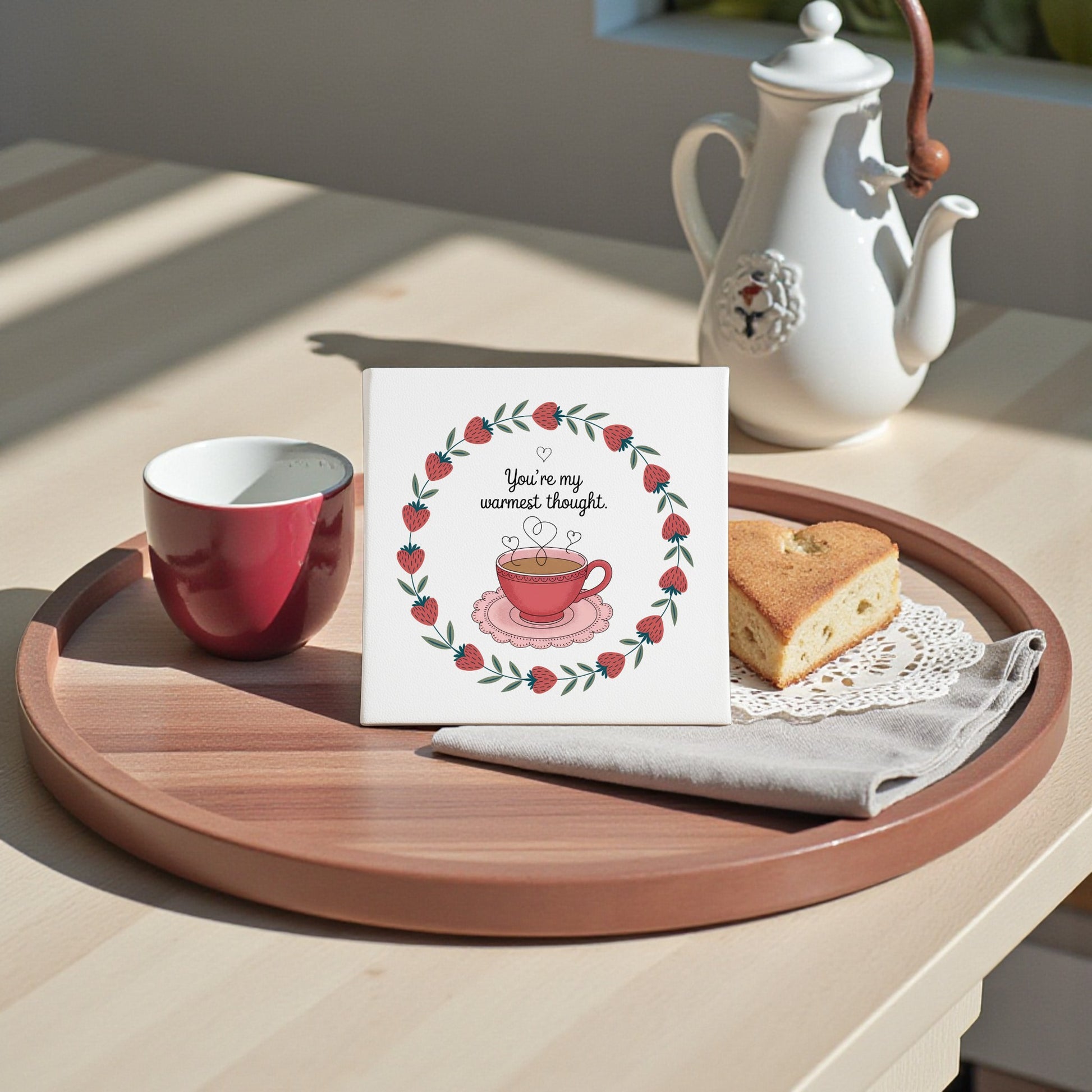 A round wooden tray displays Mirabilia Boutique's Warmest Thought Tea-Inspired Canvas Art—a square card with a teacup and "You're my warmest thought"—alongside a teapot, cup, napkin, and heart-shaped cake, ideal for tea lovers.