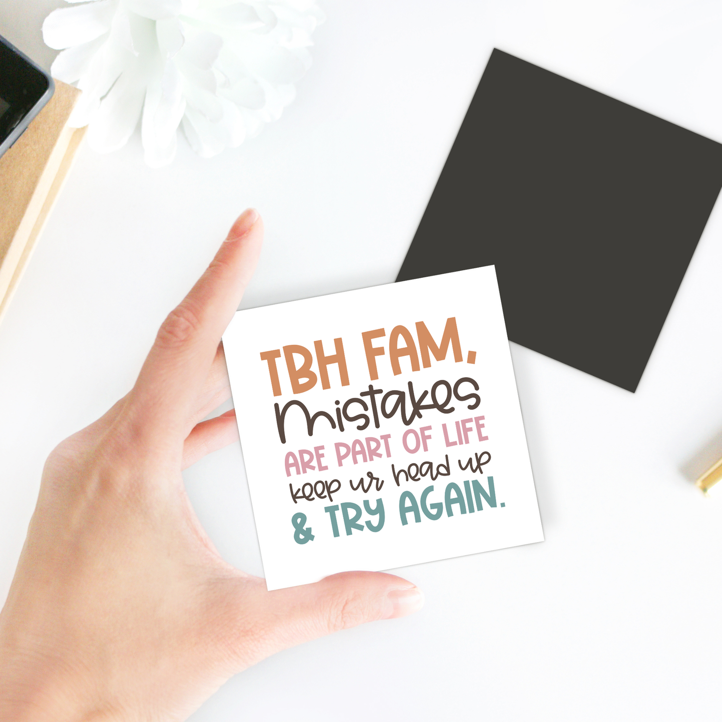 A hand holds the Mirabilia Boutique "Growth Mindset Teacher Magnet | Try Again Quote." With a white flower, black square, and gold pen on the desk, it’s perfect for motivational classroom decor or a thoughtful teacher appreciation gift.