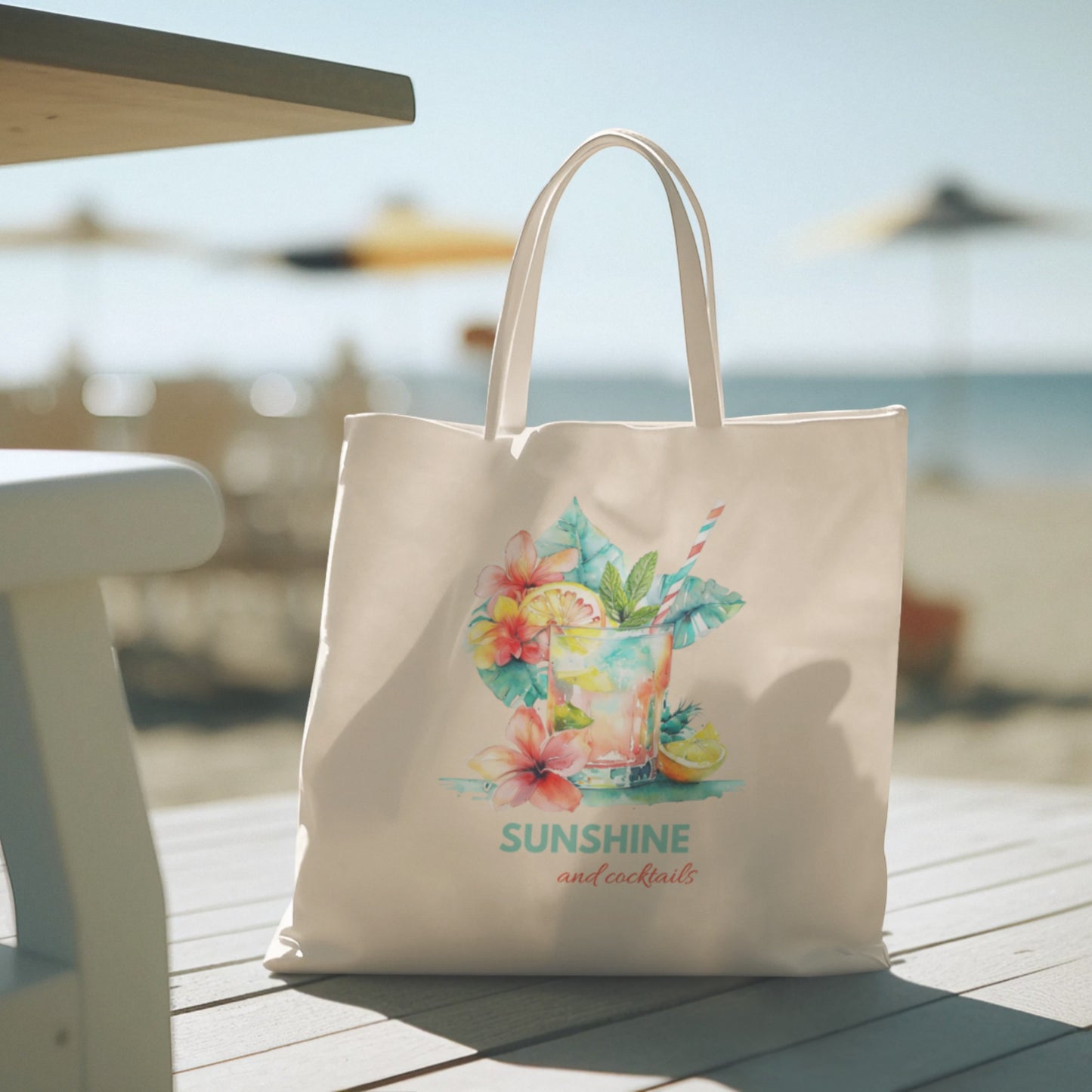 The Mirabilia Boutique "Sunshine and Cocktails Canvas Tote Bag"—featuring a vibrant beach drink, floral artwork, and the phrase "Sunshine and cocktails" on white canvas—rests on a table at a sunny beach, with chairs and umbrellas softly blurred in the background.