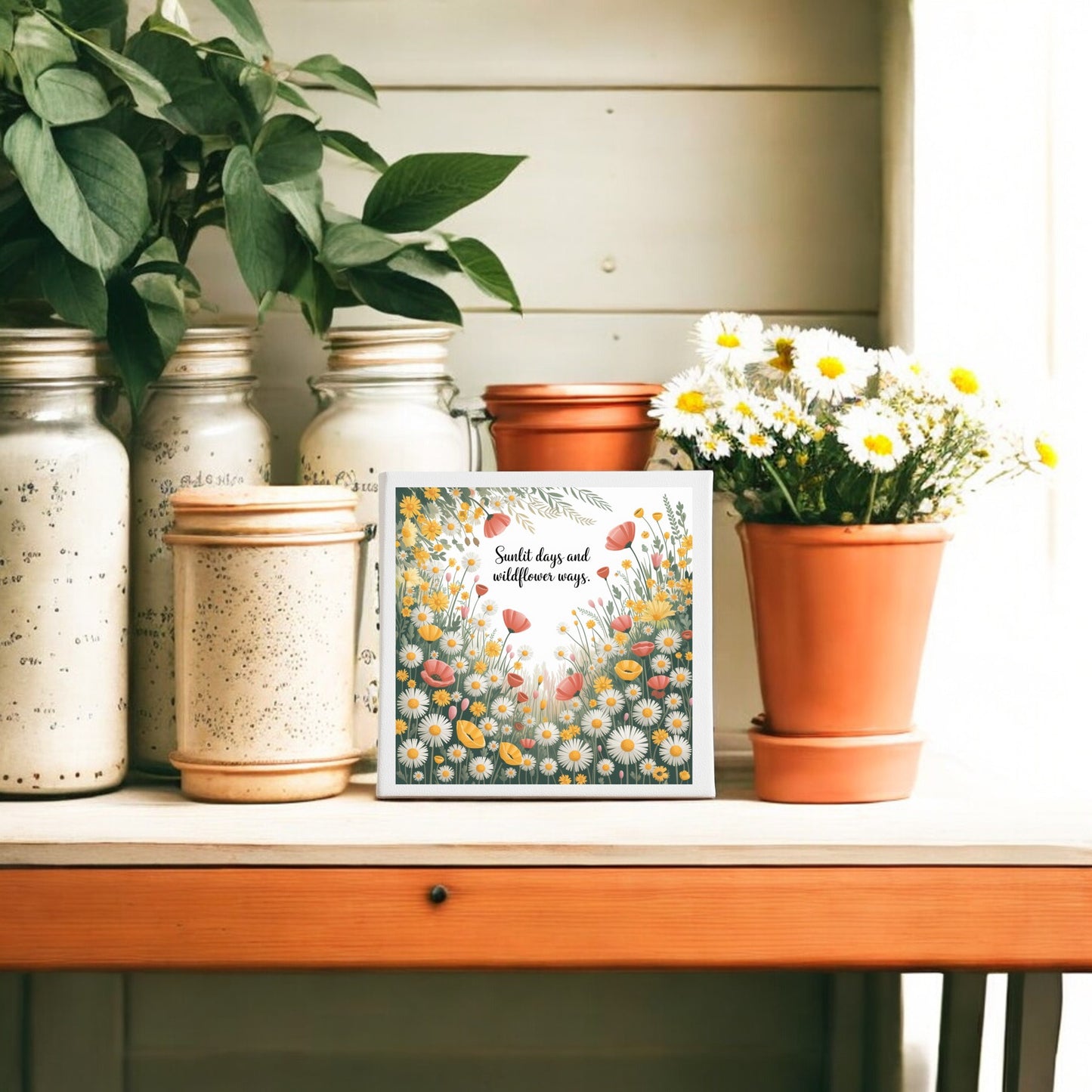A wooden table with potted daisies, green plants in jars, and Mirabilia Boutique’s Sunlit Days and Wildflower Ways 8x8 Floral Art Canvas by a bright window creates charming cottagecore decor.