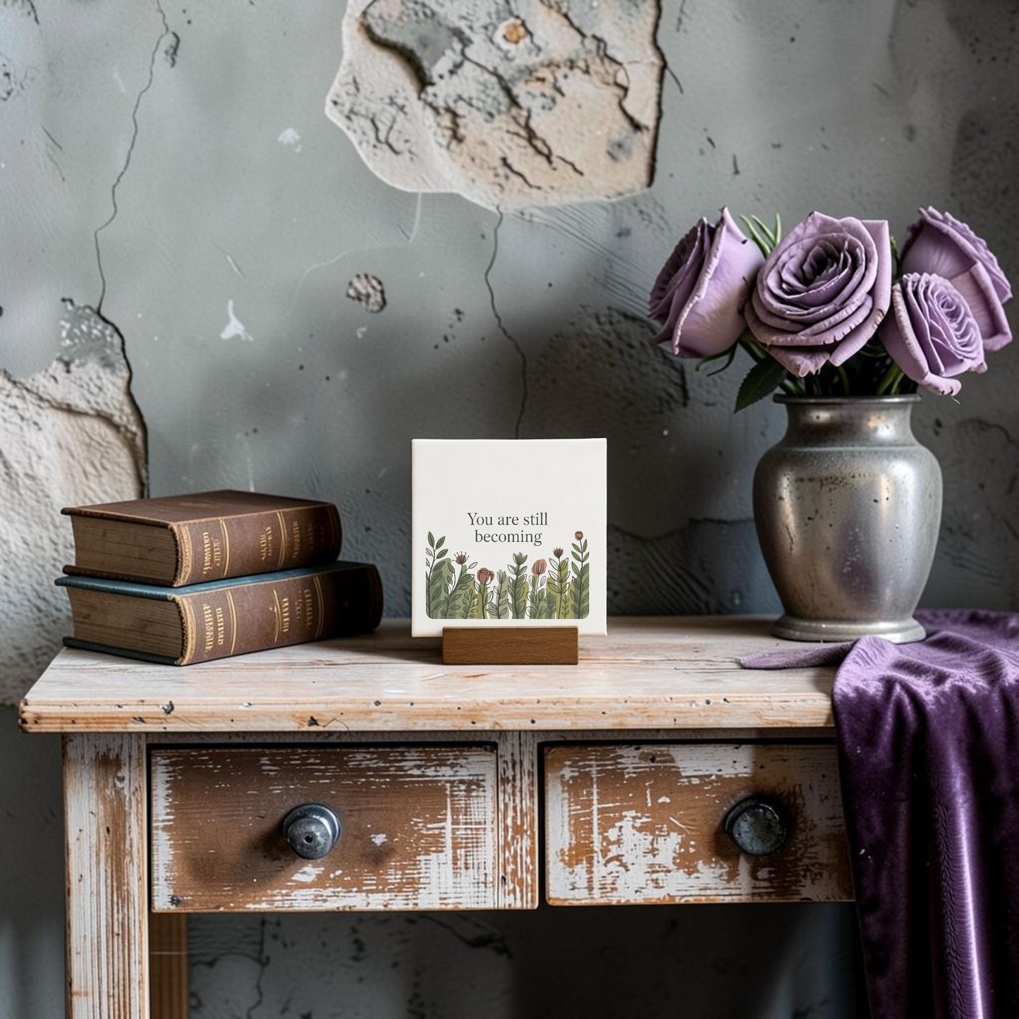 A rustic wooden table displays stacked books, a Mirabilia Boutique "You Are Still Becoming" mini canvas sign with floral art, a silver vase of purple roses, and a draped purple cloth, all set against a distressed wall.