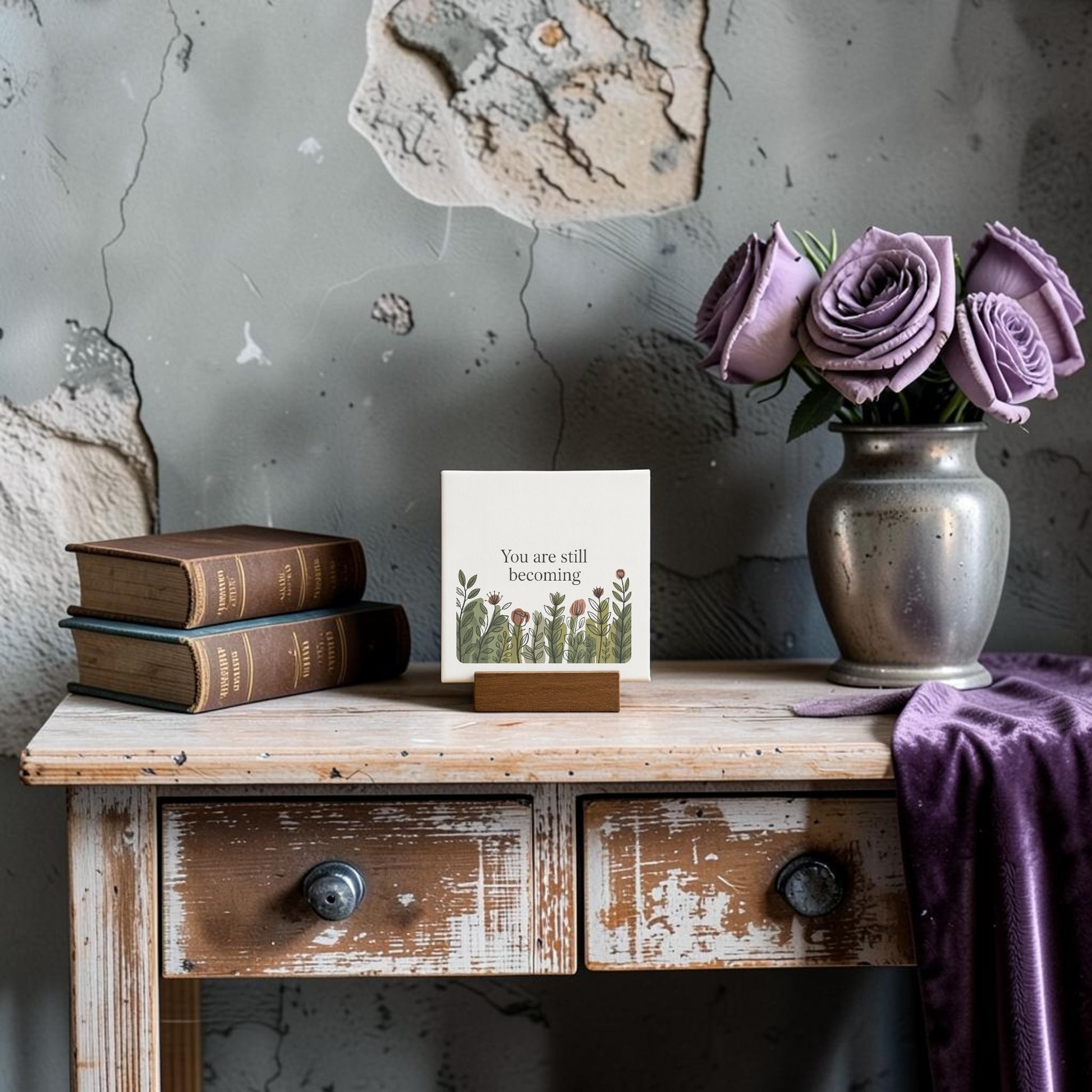 A rustic wooden table displays stacked books, a Mirabilia Boutique "You Are Still Becoming" mini canvas sign with floral art, a silver vase of purple roses, and a draped purple cloth, all set against a distressed wall.