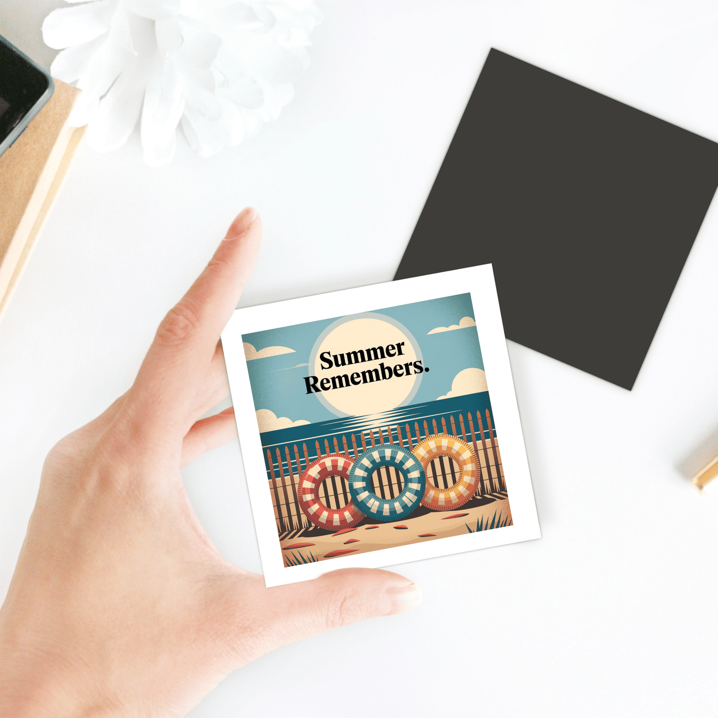 A hand holds the "Summer Remembers Magnet – Vintage Coastal Fridge Art" by Mirabilia Boutique, featuring colorful lifebuoys spelling "COOL"—ideal coastal decor or a beach lover gift. A flower and black square accent sit on the white table.