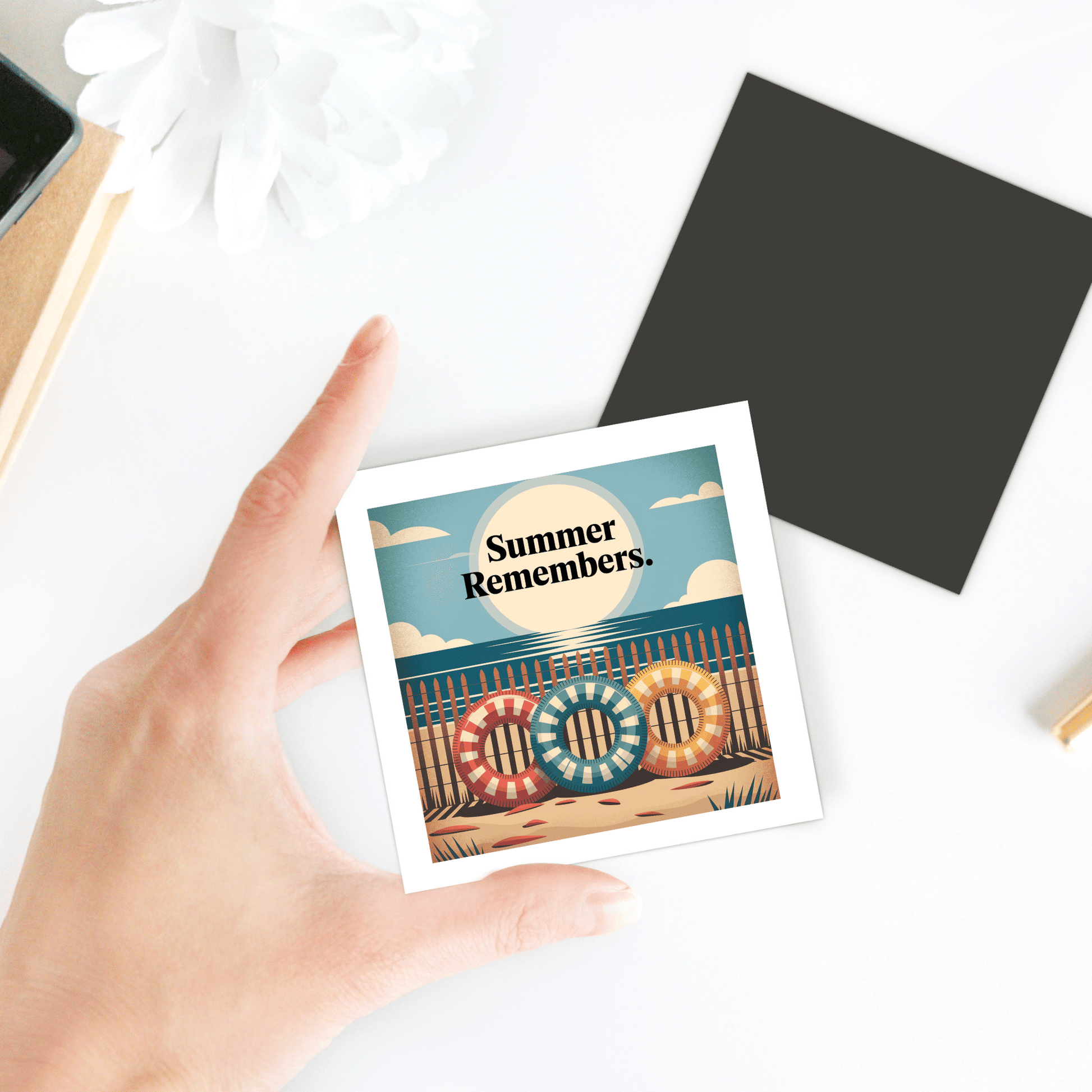 A hand holds the "Summer Remembers Magnet – Vintage Coastal Fridge Art" by Mirabilia Boutique, featuring colorful lifebuoys spelling "COOL"—ideal coastal decor or a beach lover gift. A flower and black square accent sit on the white table.