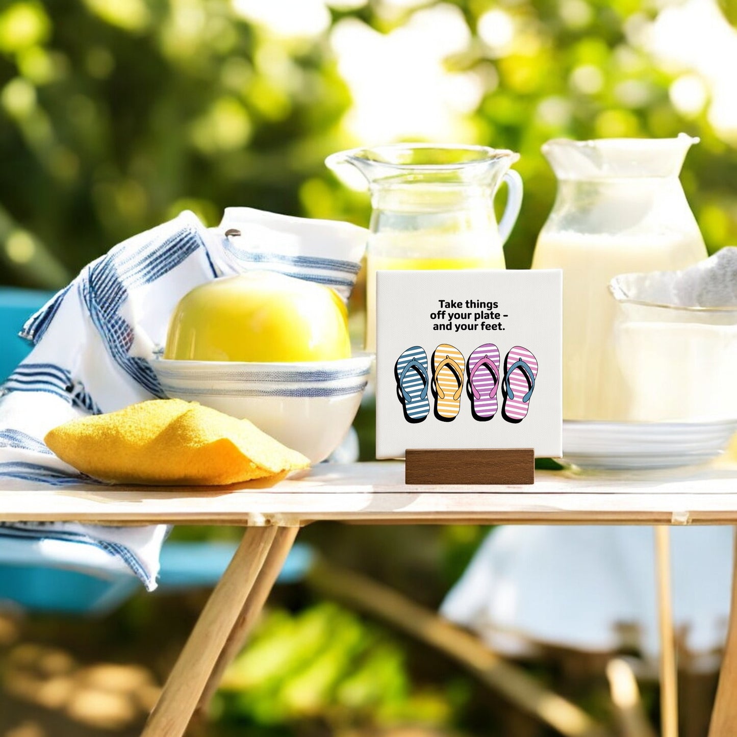 On an outdoor table sits a striped towel, a bowl with a green apple, milk pitchers, and Mirabilia Boutique’s "Take Things Off Your Feet" 4x4 mini canvas sign—perfect for summer decor and relaxation.