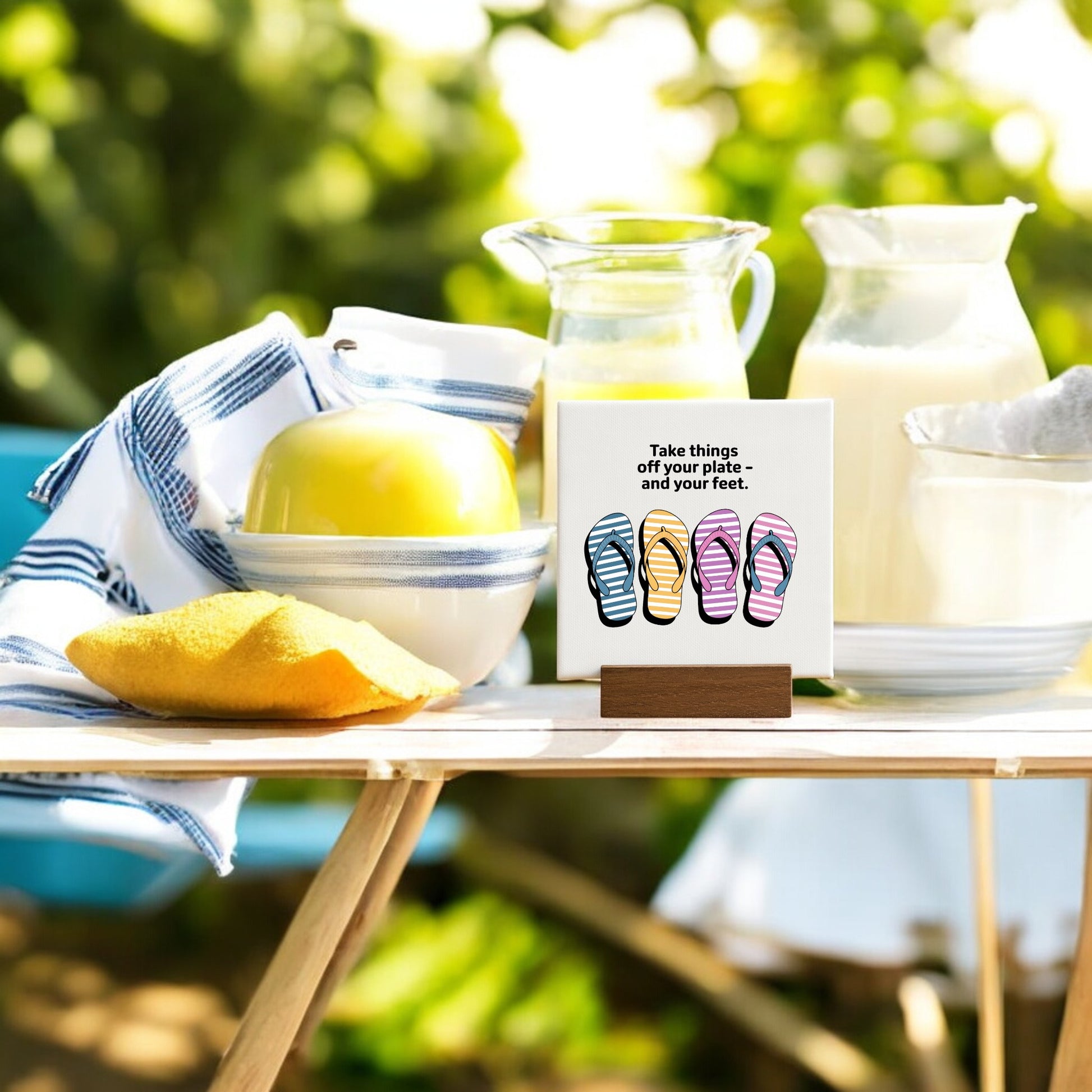 On an outdoor table sits a striped towel, a bowl with a green apple, milk pitchers, and Mirabilia Boutique’s "Take Things Off Your Feet" 4x4 mini canvas sign—perfect for summer decor and relaxation.