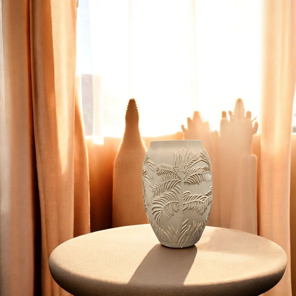 The Sand Palm Desert Vase for Summer Decor by Mirabilia Boutique features a palm leaf pattern and sits on a round table before sunlit peach curtains and a window, adding desert-inspired style. Blurred shapes in the background suggest plants or decor.