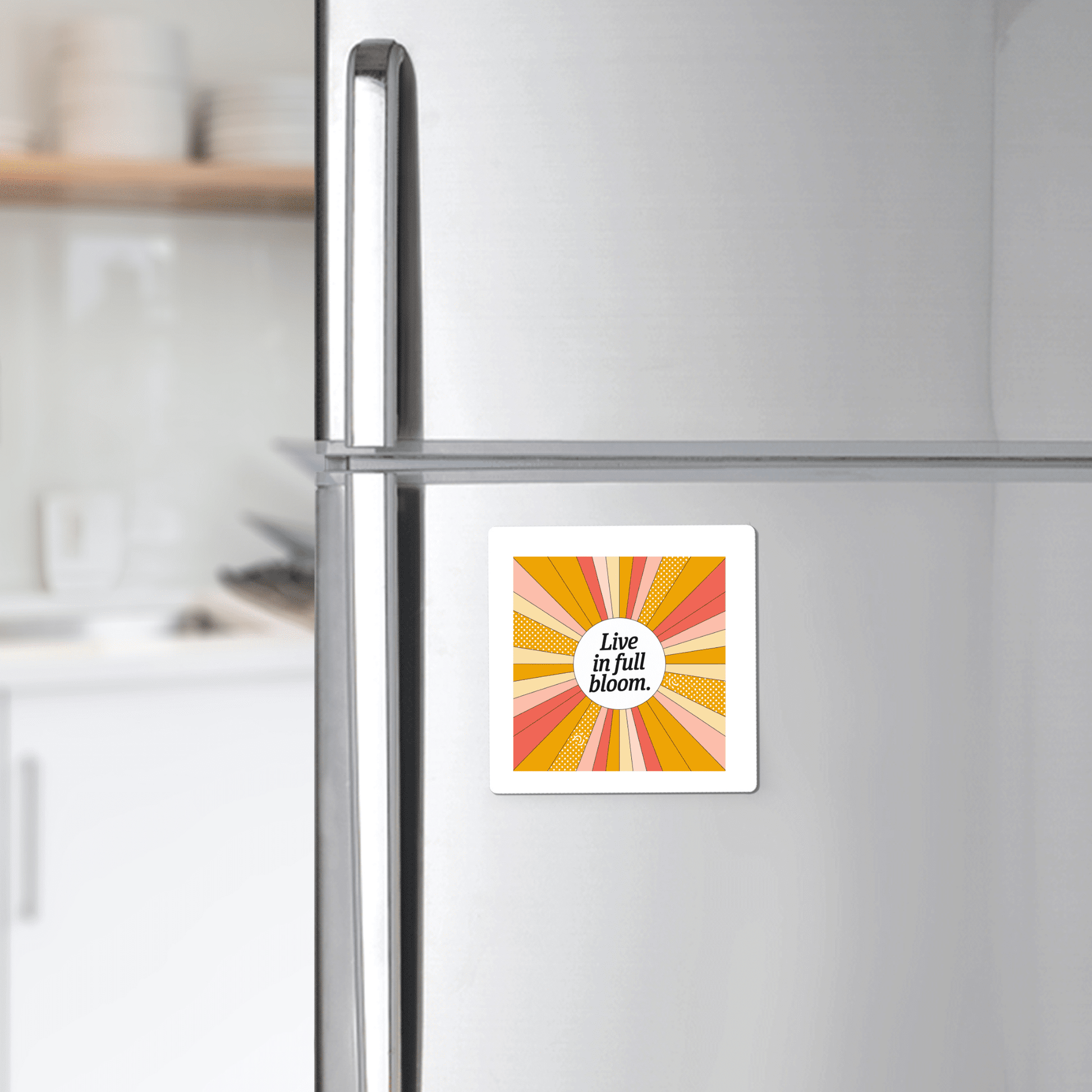 A silver refrigerator displays the Live in Full Bloom Magnet – Summer Sunburst Mini Art by Mirabilia Boutique. The vibrant square, with sunburst stripes in yellow, orange, and pink, stands out against a softly blurred kitchen backdrop.