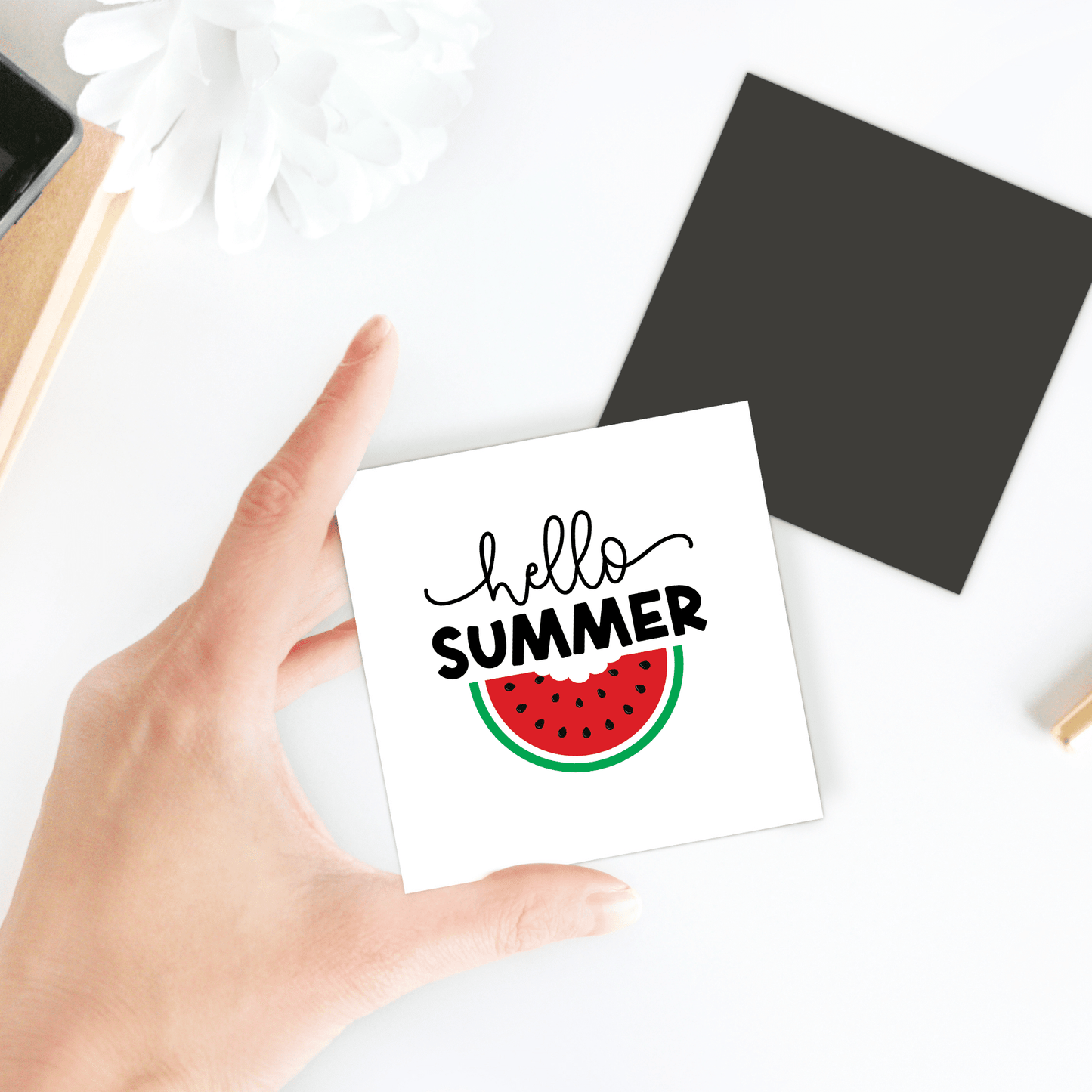 A hand holds Mirabilia Boutique’s Hello Summer Watermelon Magnet – Juicy Seasonal Fridge Art, featuring "hello SUMMER" above a watermelon slice––ideal as summer decor for your fridge. Black square, white flower, and objects rest on the white table.