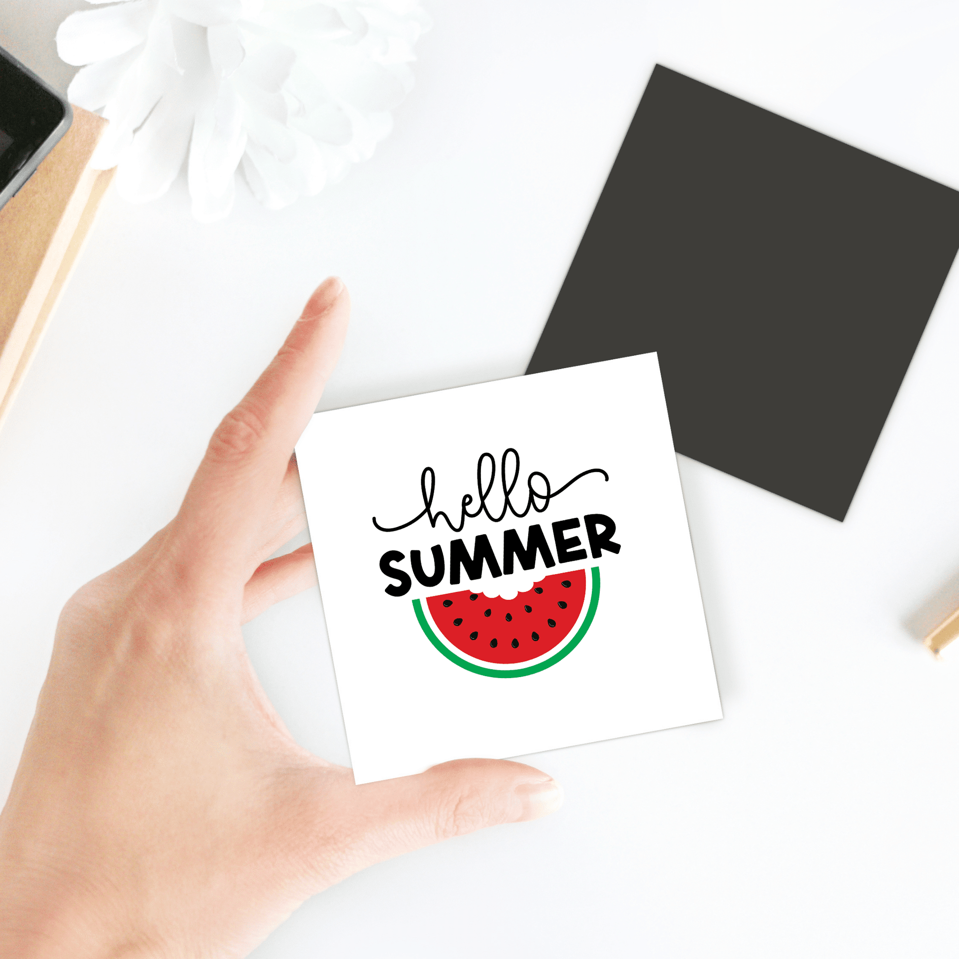 A hand holds Mirabilia Boutique’s Hello Summer Watermelon Magnet – Juicy Seasonal Fridge Art, featuring "hello SUMMER" above a watermelon slice––ideal as summer decor for your fridge. Black square, white flower, and objects rest on the white table.