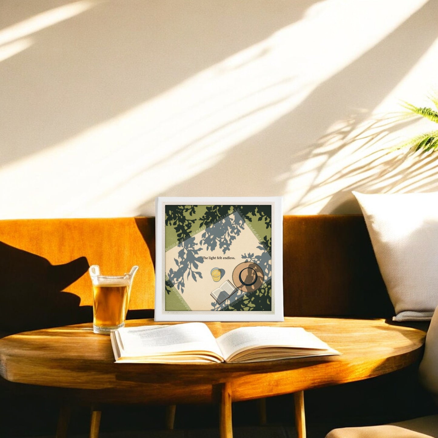 A sunlit seating area features Mirabilia Boutique's "The Light Felt Endless Canvas – Summer Stillness Art" (8x8), displayed on a wooden table with an open book, iced coffee, and leafy shadows, set by a mustard couch against a cream wall.