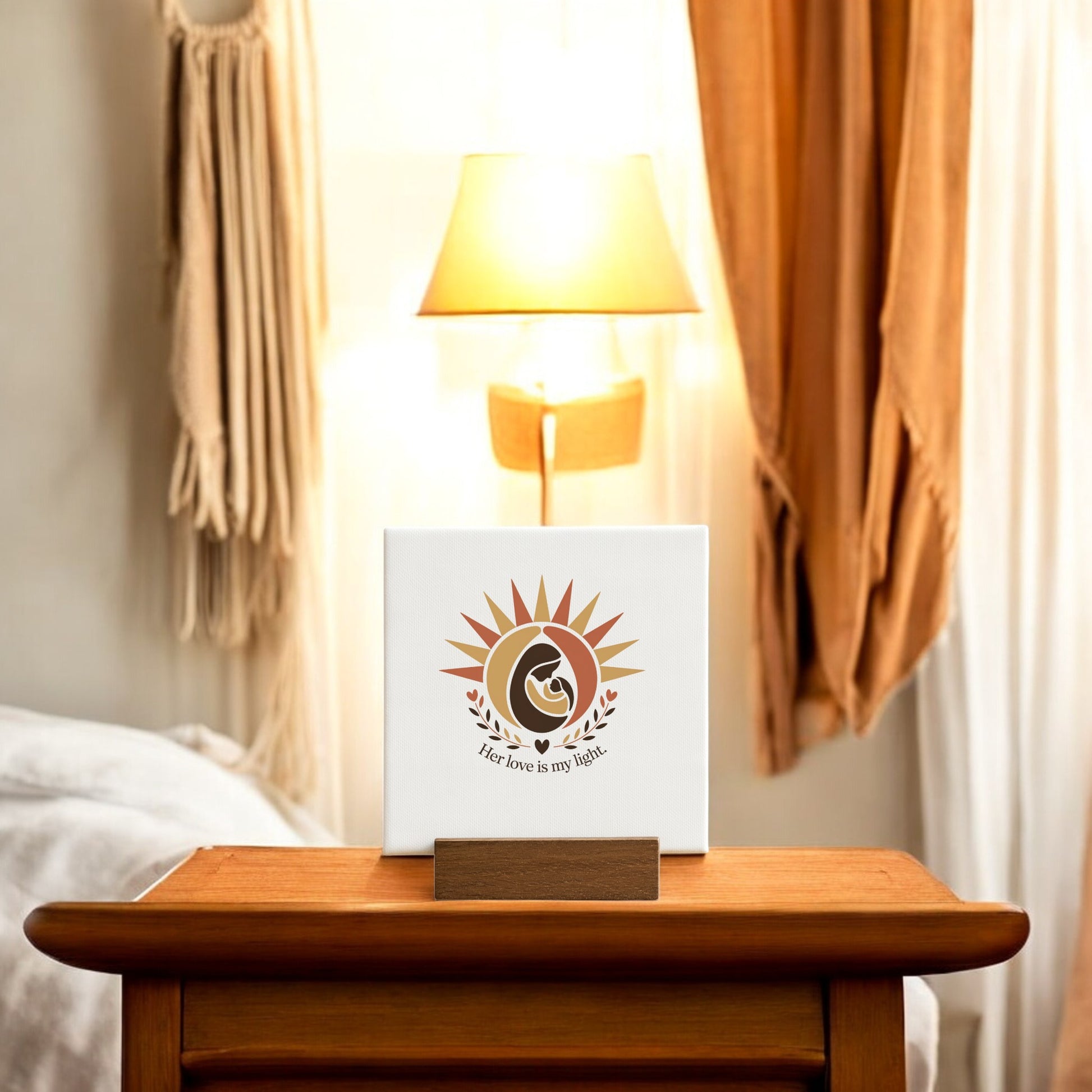 A wooden nightstand displays the Mirabilia Boutique "Her Love Is My Light" Mini Sign – Mother’s Day Canvas, featuring a sun and leaf design, making a heartfelt keepsake for mom. A lit lamp, bed, and beige curtains appear softly in the background.