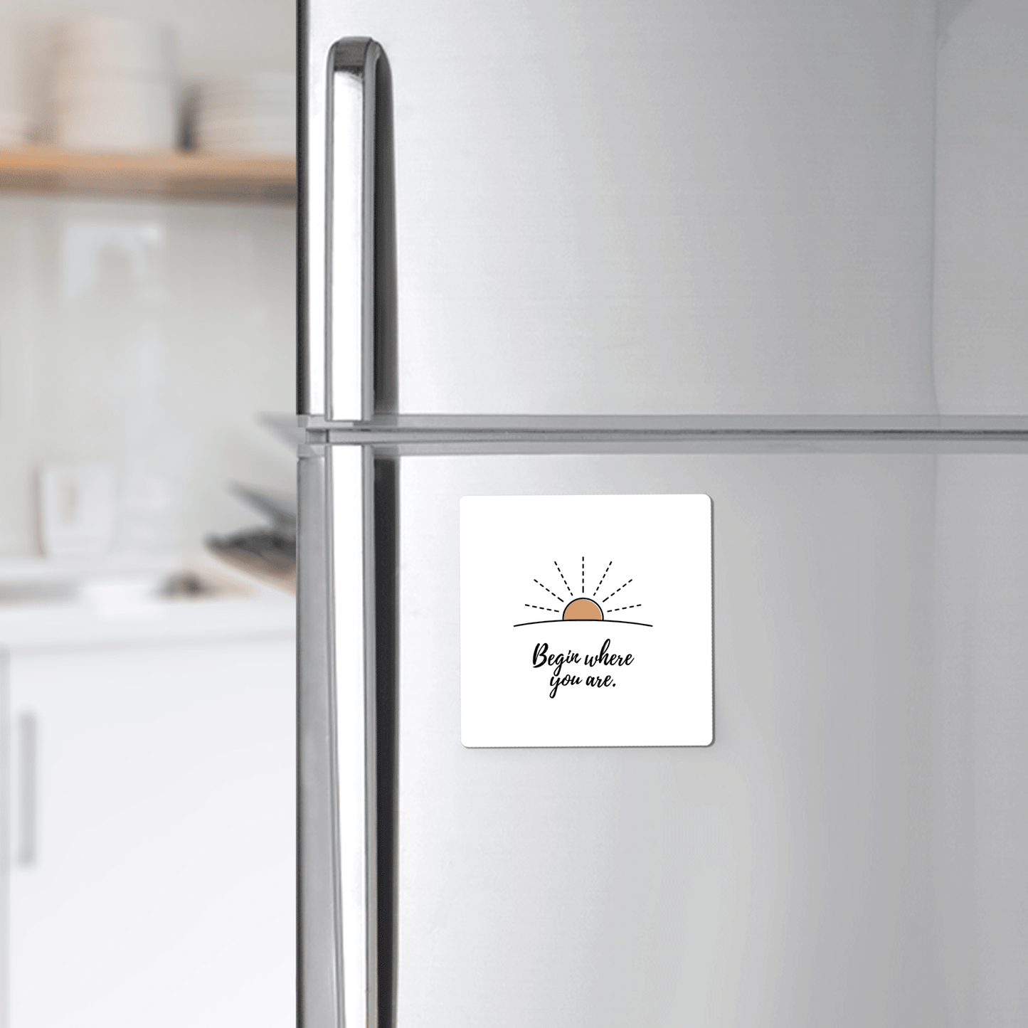 A silver fridge displays Mirabilia Boutique’s “Begin Where You Are Magnet – Sunrise Affirmation,” featuring a simple sun sketch and handwritten text. In the background, a bright kitchen adds to the cheerful vibe.