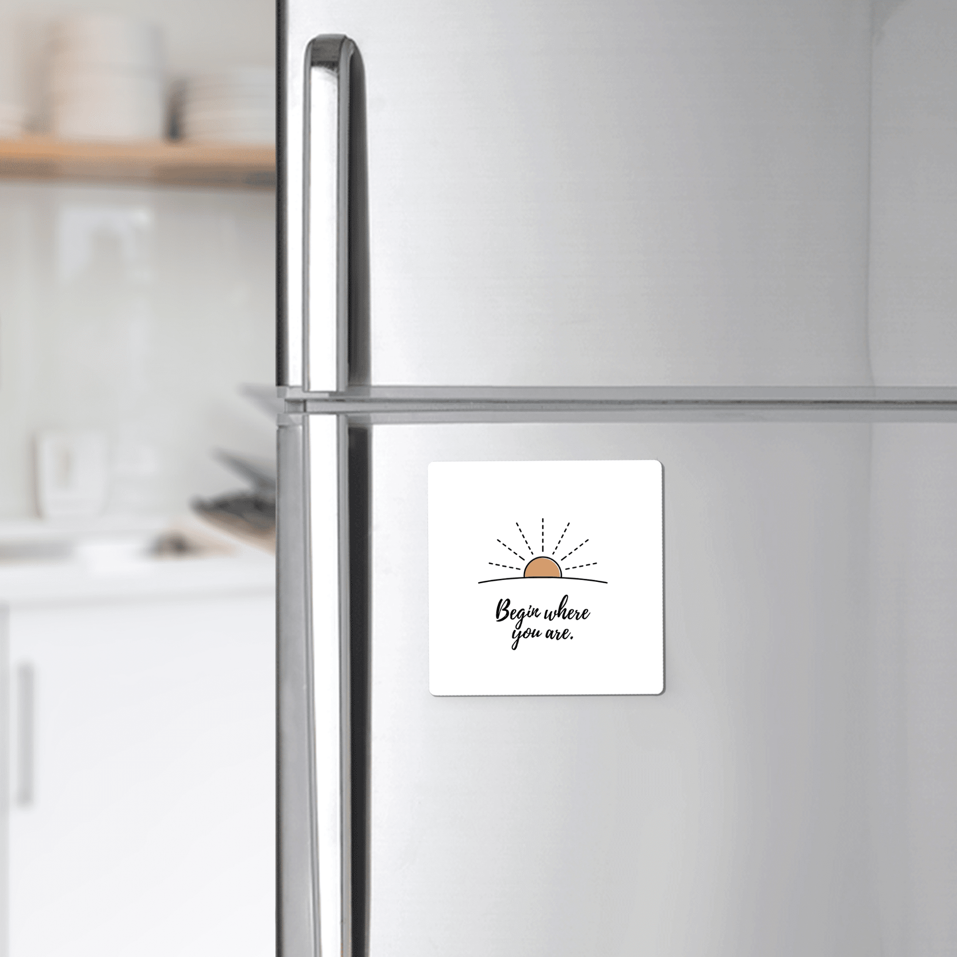 A silver fridge displays Mirabilia Boutique’s “Begin Where You Are Magnet – Sunrise Affirmation,” featuring a simple sun sketch and handwritten text. In the background, a bright kitchen adds to the cheerful vibe.
