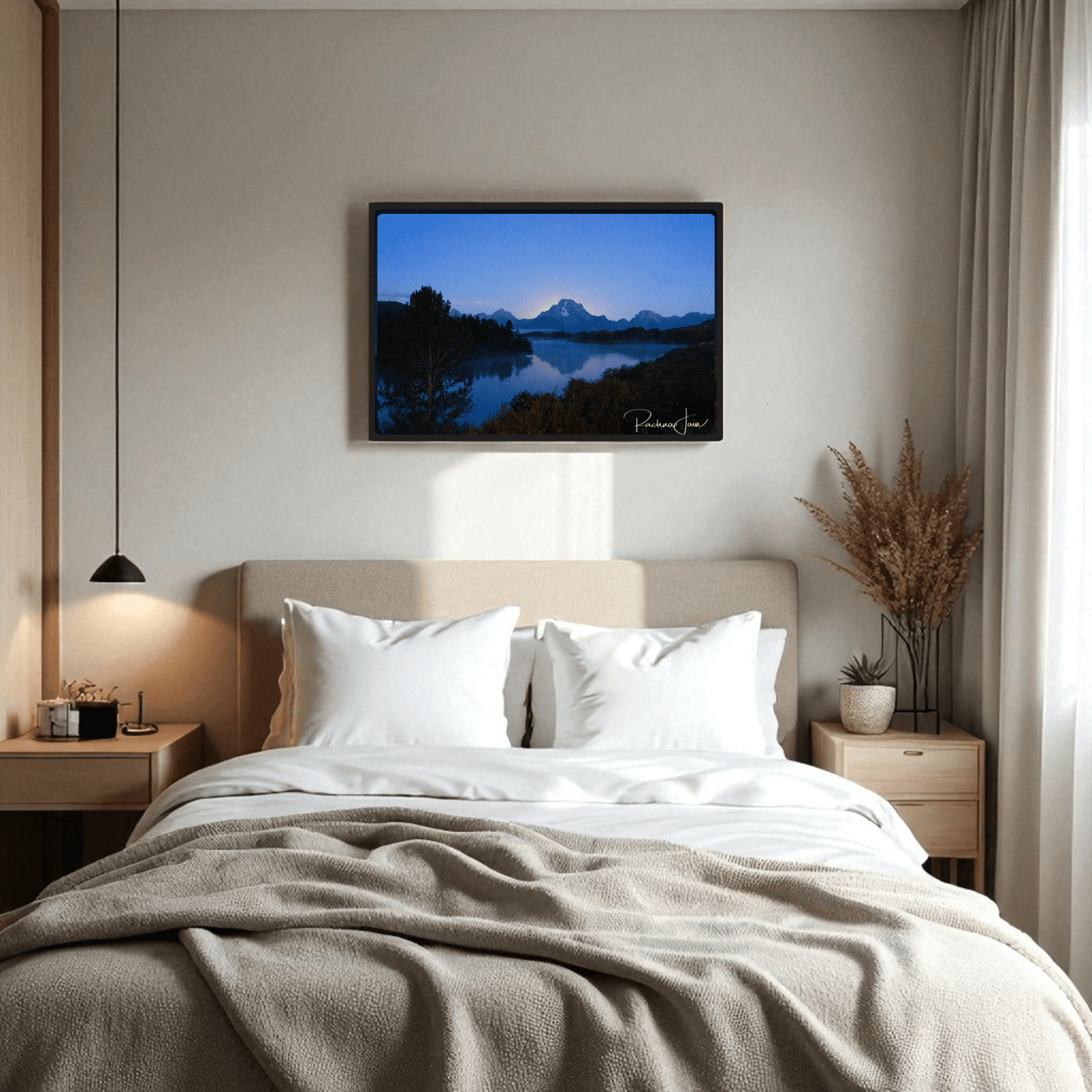 A cozy bed with white pillows is centered in a modern room. Above it hangs the “Sunrise at Oxbow Bend” framed Grand Teton art print by Rachna Jain from Mirabilia Boutique. Light wood accents and dried plants add warmth to the serene space.