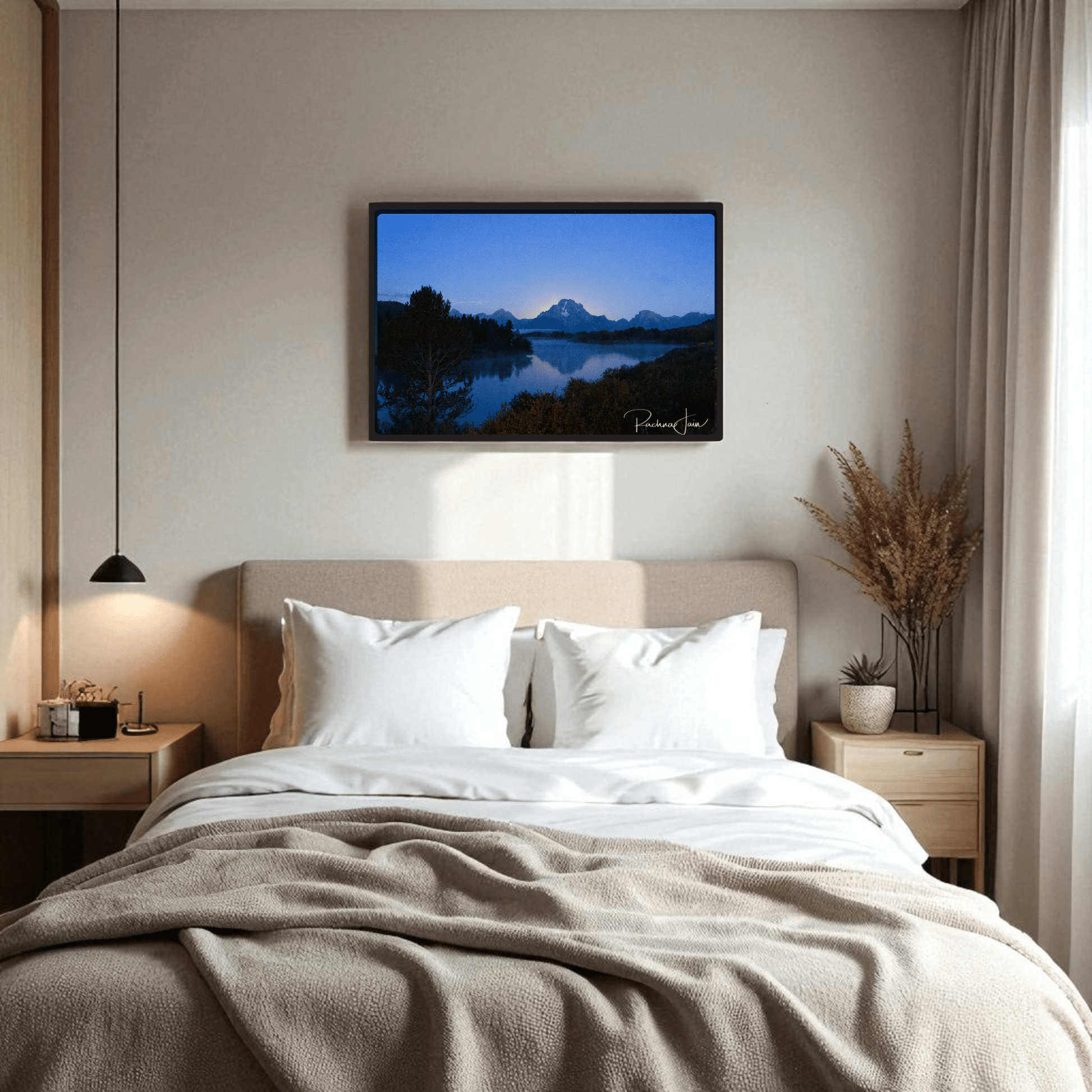 A cozy bed with white pillows is centered in a modern room. Above it hangs the “Sunrise at Oxbow Bend” framed Grand Teton art print by Rachna Jain from Mirabilia Boutique. Light wood accents and dried plants add warmth to the serene space.