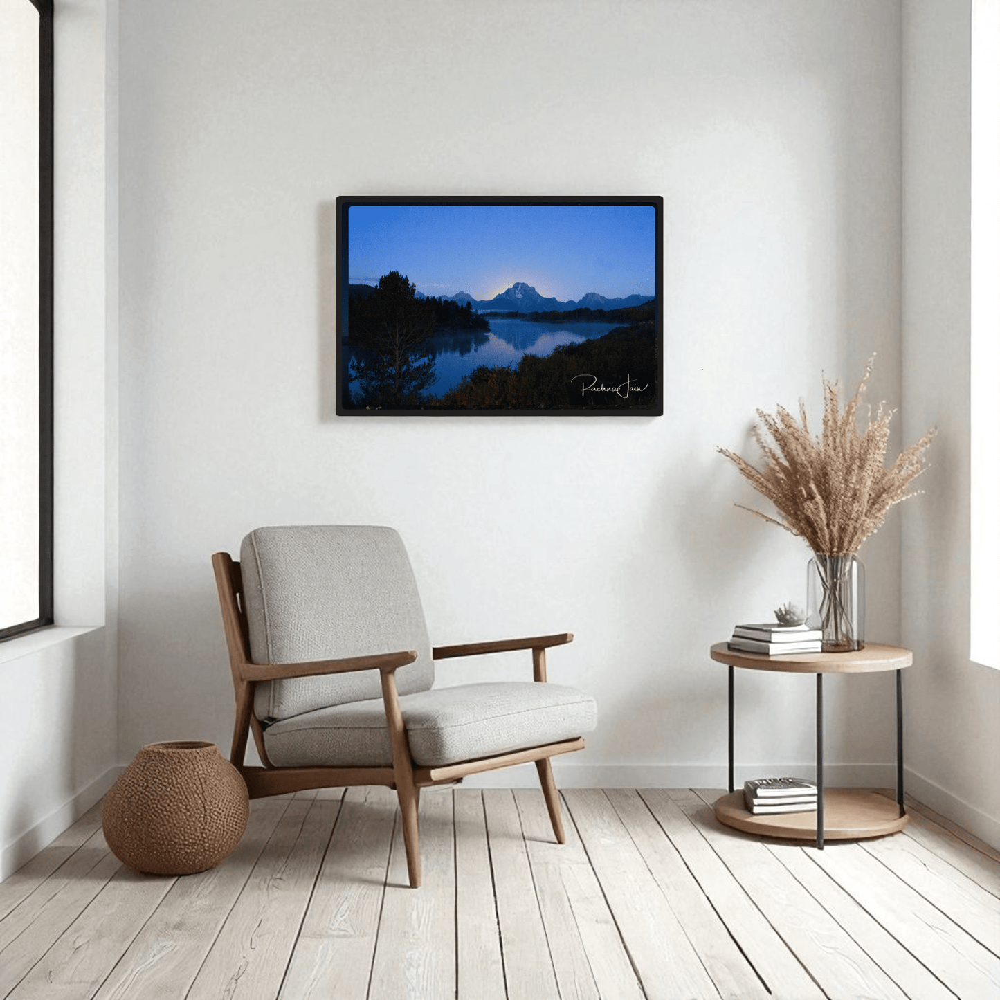 A modern living room features a light gray armchair, round side table with dried grasses, a woven basket, and "Sunrise at Oxbow Bend"—a framed Grand Teton art print by Rachna Jain from Mirabilia Boutique—on a white wall.